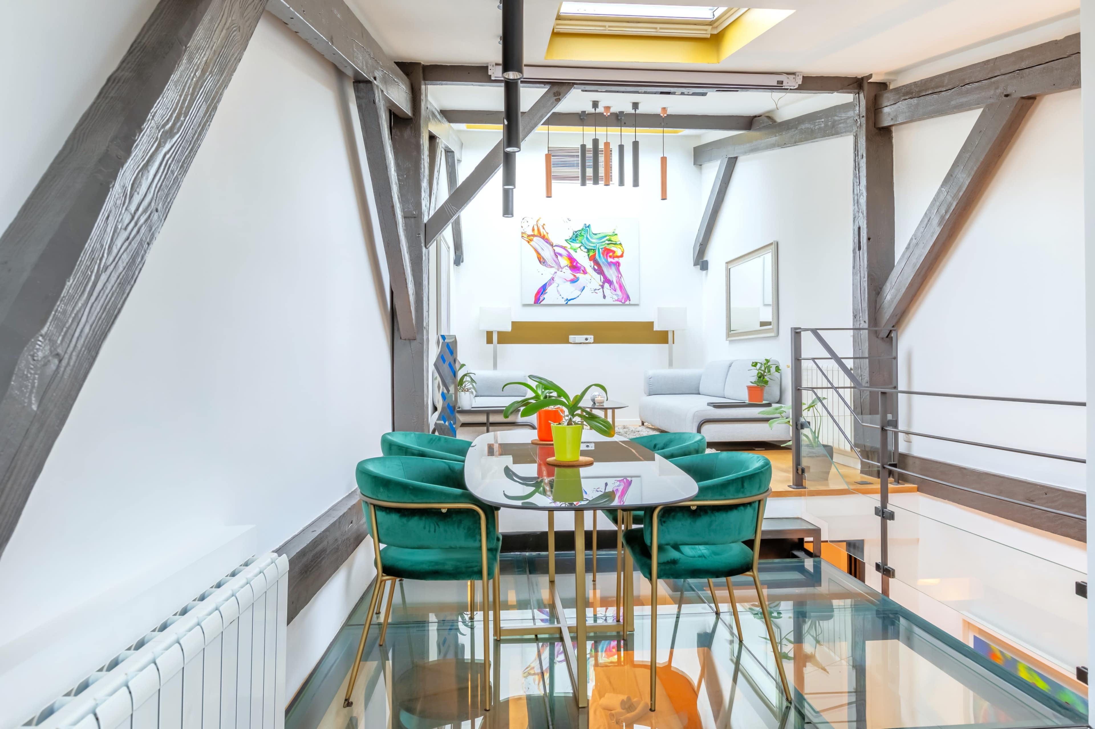 The image shows a modern interior space featuring a glass-top dining table with green chairs, wooden beams, and a living area visible in the background.