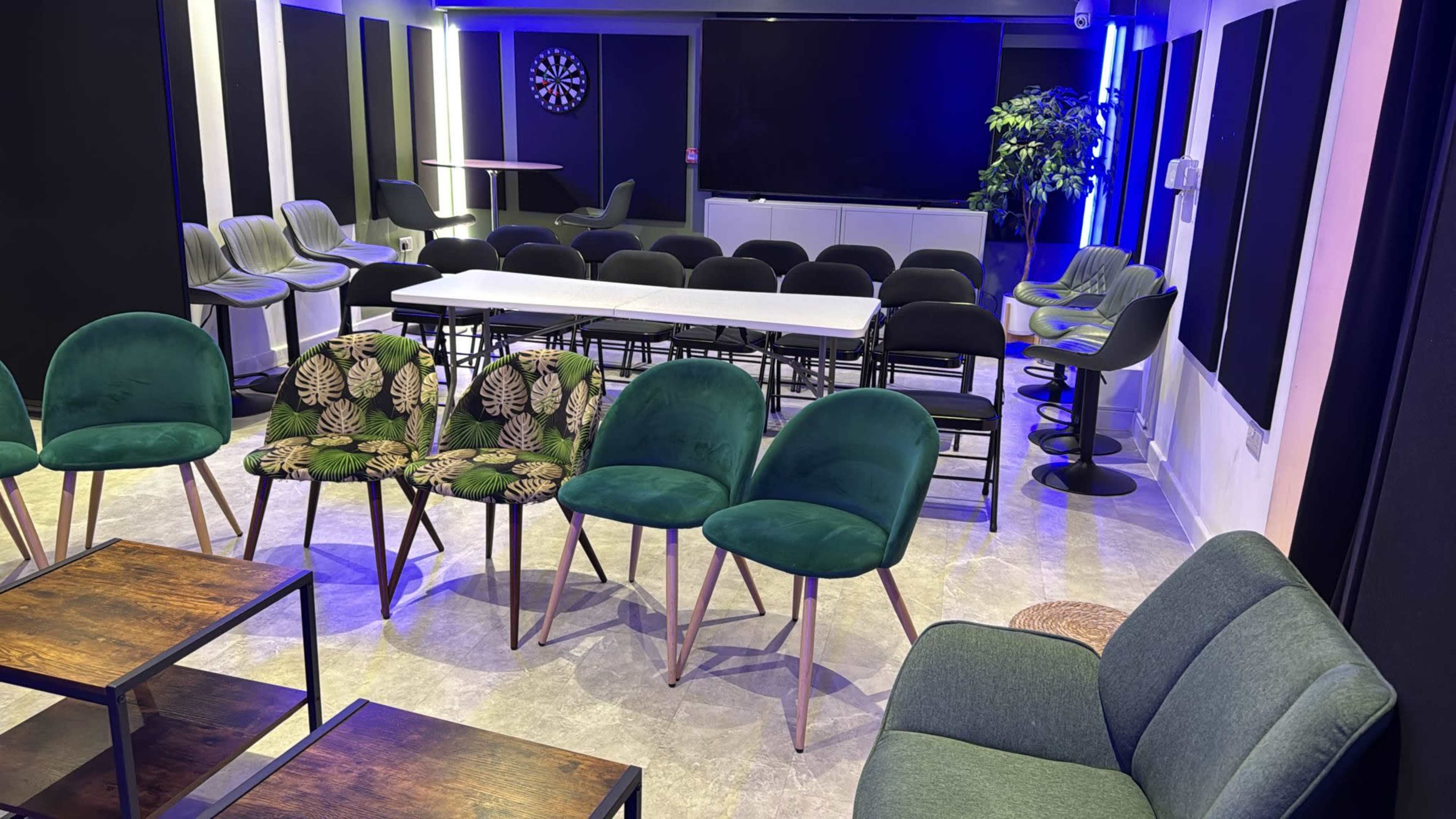 The image shows a modern meeting space with a variety of seating arrangements, including chairs, tables, and a dartboard on the wall.