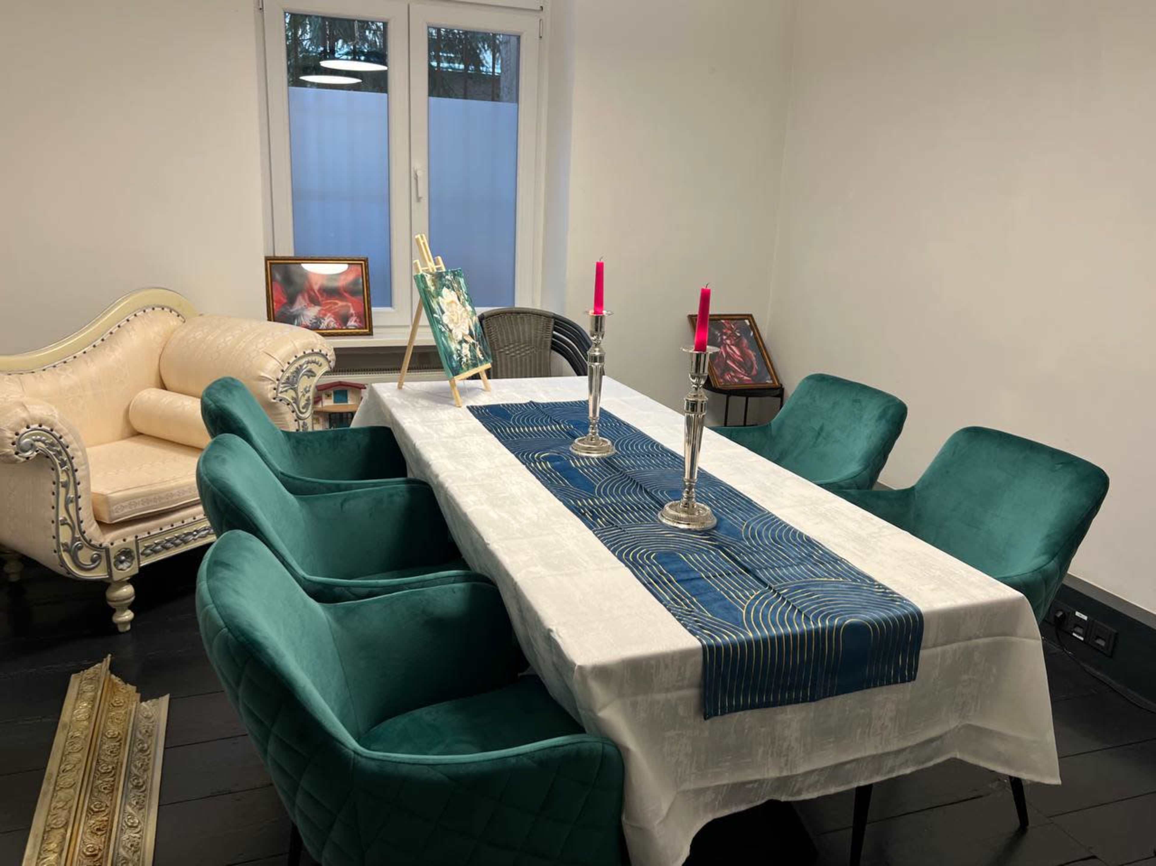 A dining room features a white tablecloth over a long table surrounded by green chairs, with a decorative runner and candles, alongside a cream-colored sofa and framed artwork on the walls.