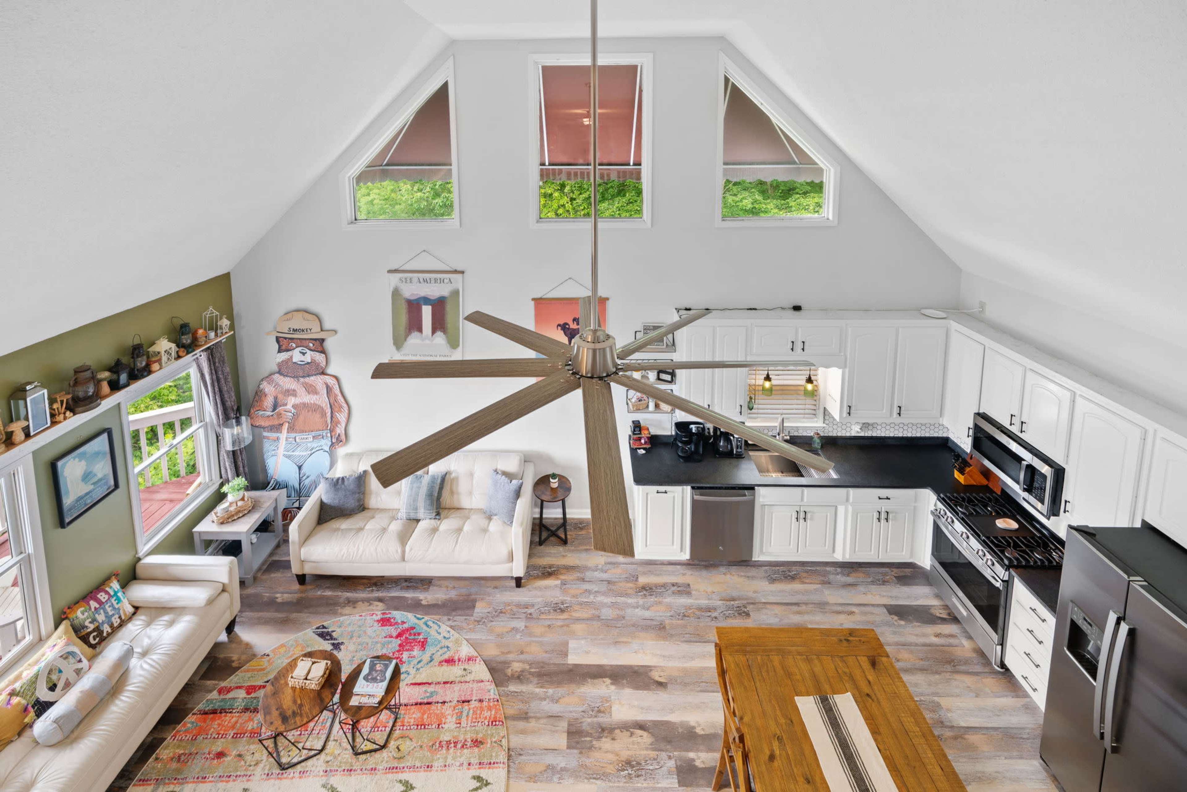 The image shows a spacious kitchen and living area with a large ceiling fan, white cabinetry, and a colorful area rug.