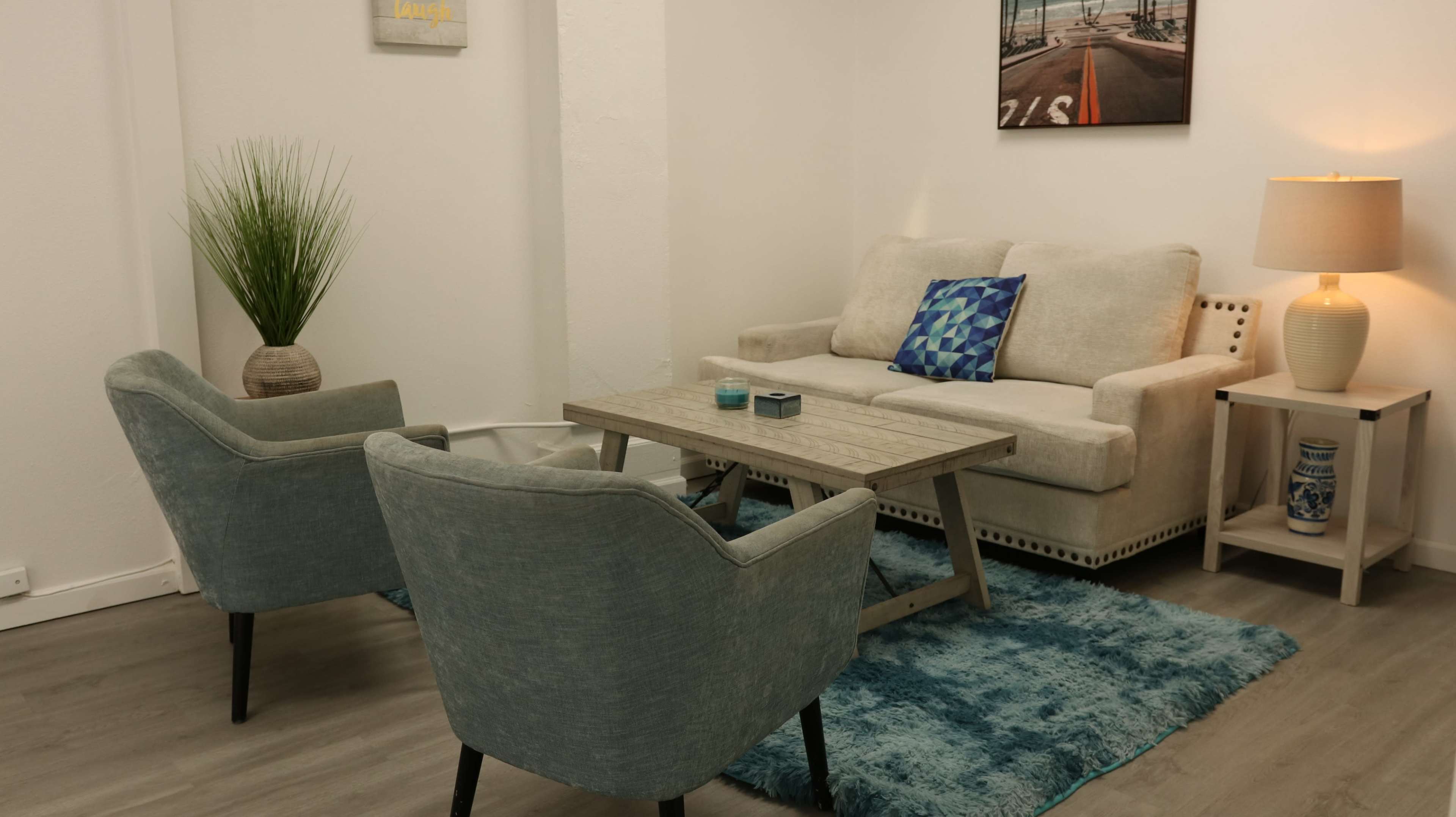 The image depicts a small, modern living area featuring a beige sofa, two light blue armchairs, a wooden coffee table, and a decorative rug.