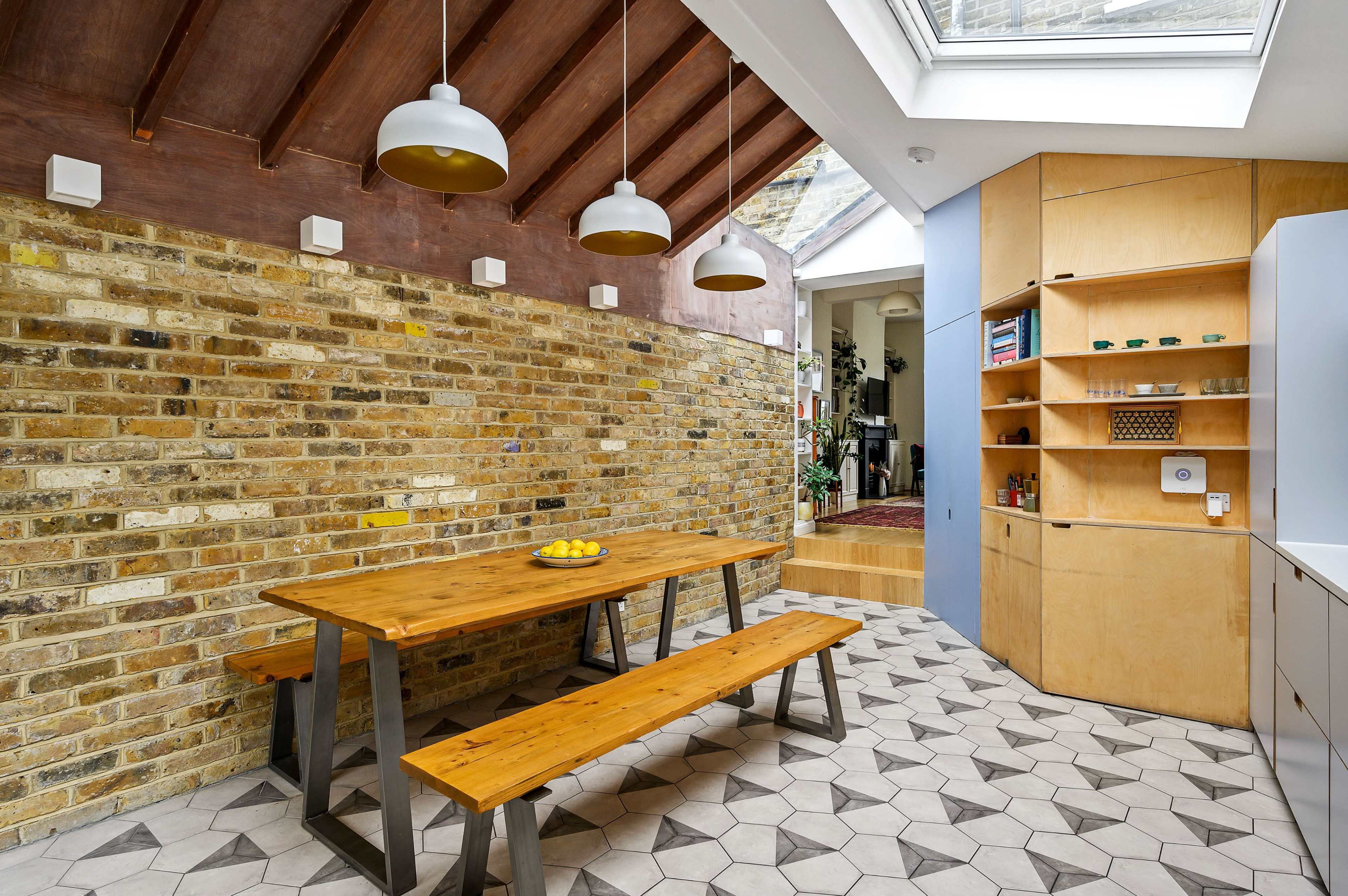 The image depicts a modern kitchen with a wooden table and benches, a textured brick wall, and a skylight illuminating the space.