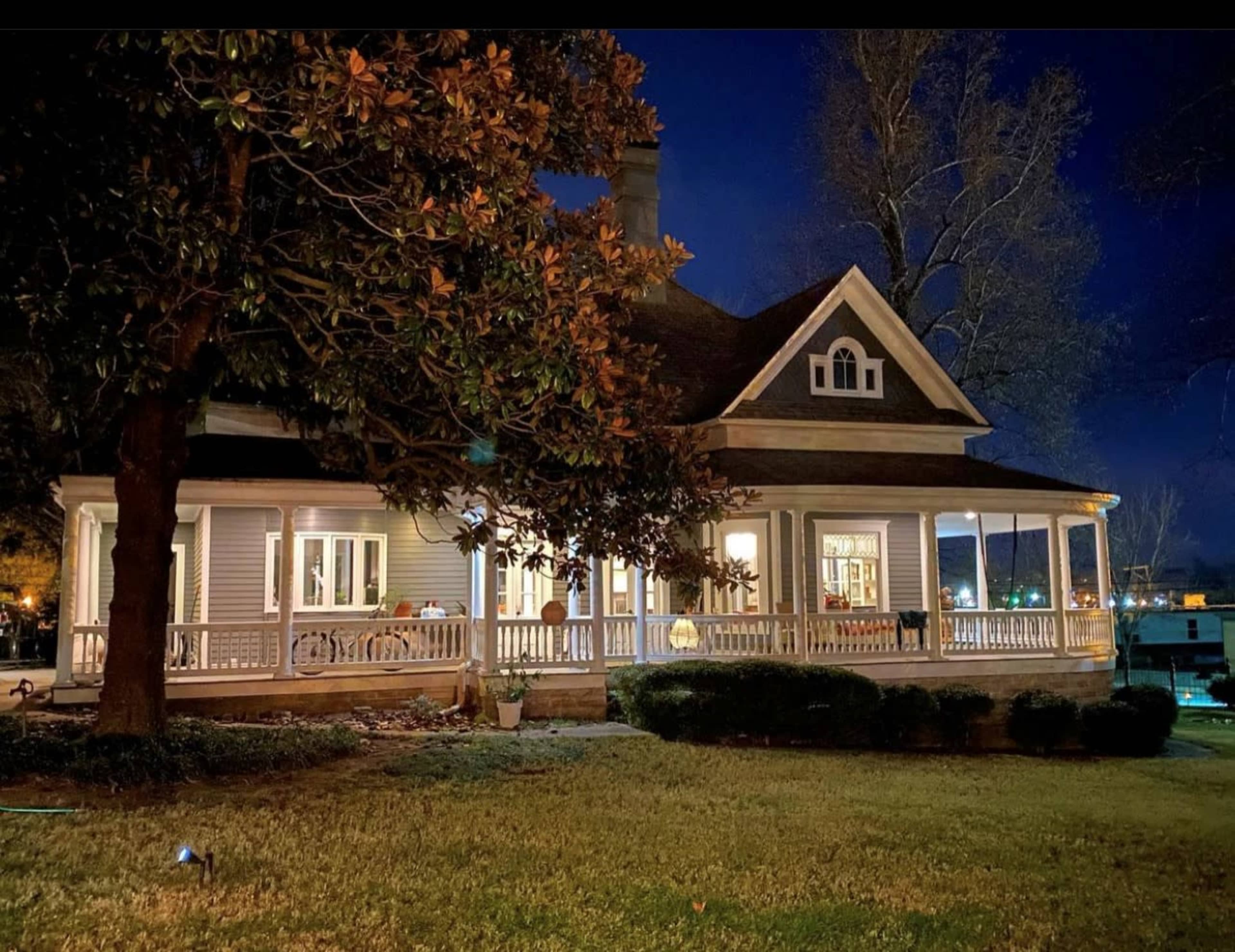 A two-story house with a wraparound porch and large windows is illuminated at night, surrounded by grass and trees.