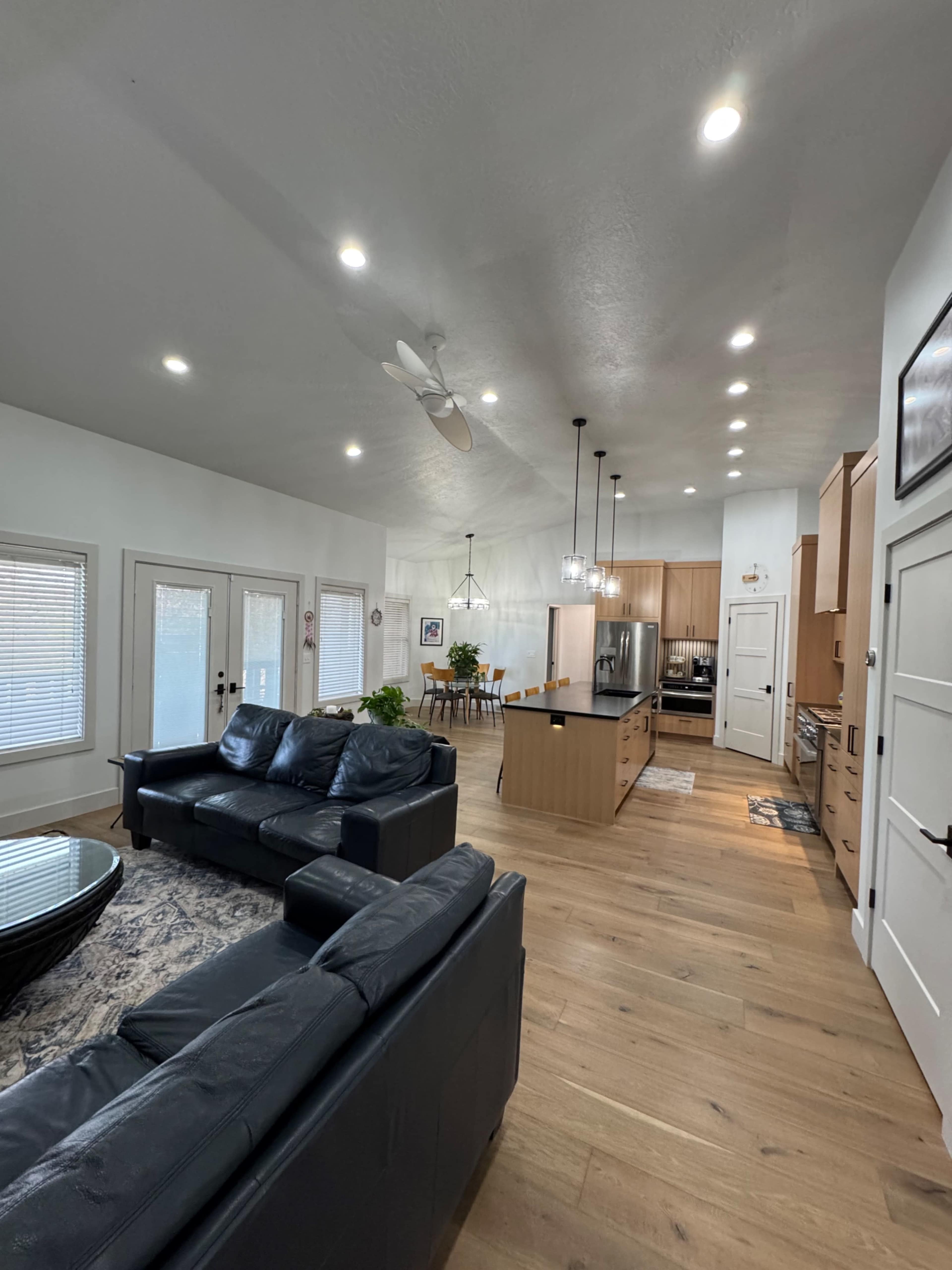 The image shows a modern, open-concept living area with two black sofas facing a kitchen equipped with wooden cabinetry and stainless steel appliances.