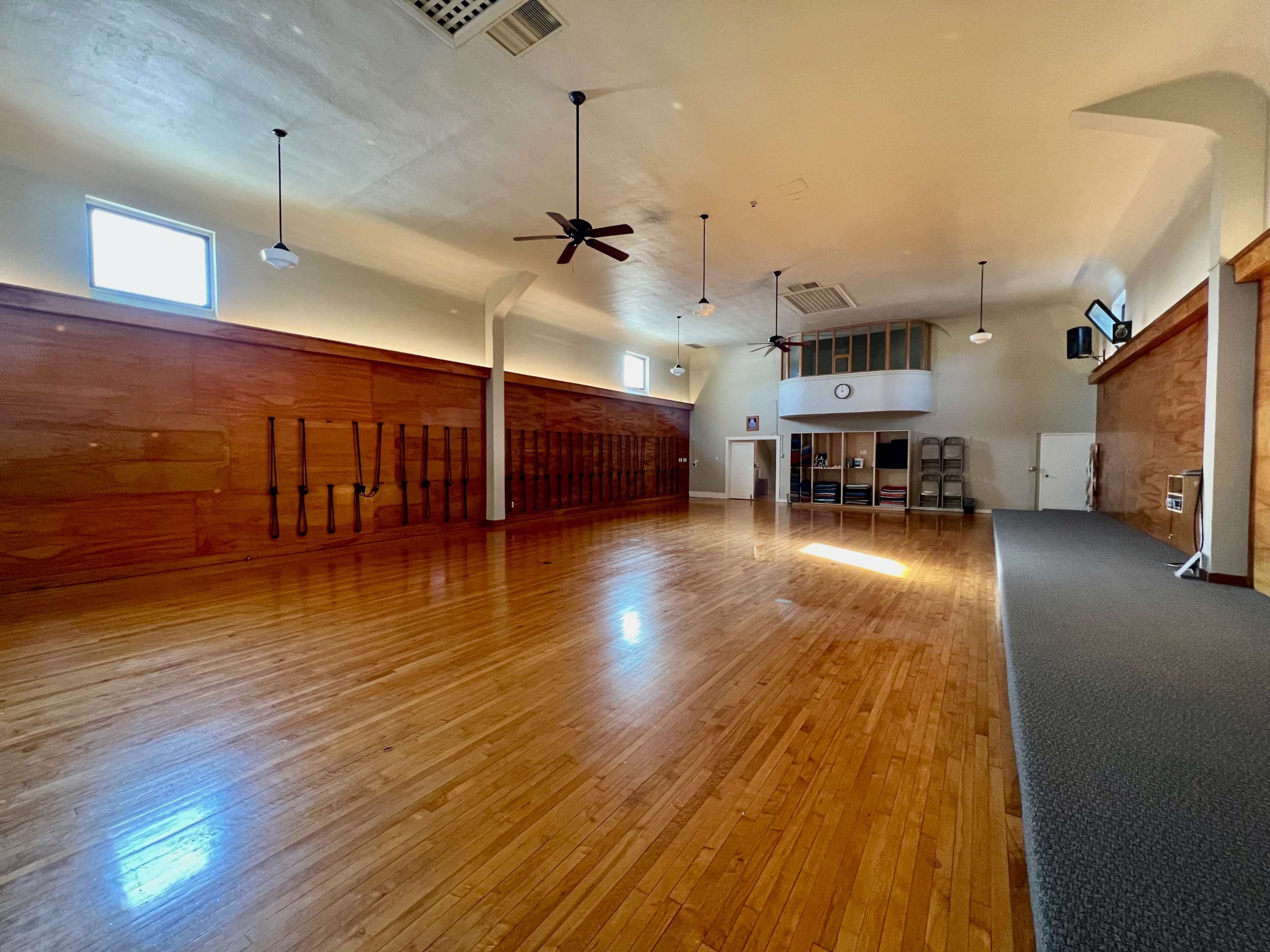 A spacious, empty room with polished wooden floors, ceiling fans, and large windows, equipped with wooden walls and some chairs stacked against one side.