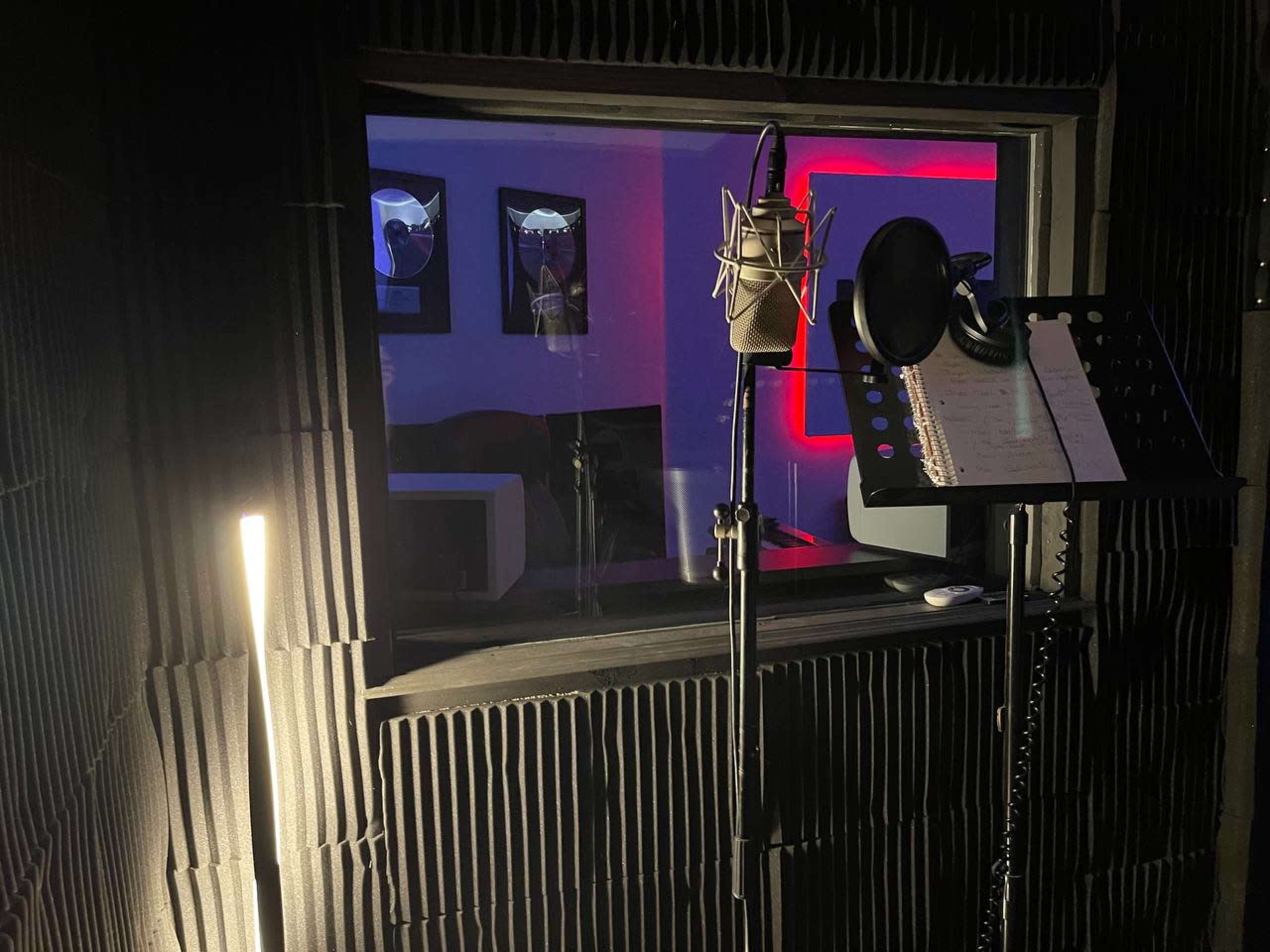 A soundproof recording booth with a microphone, music stand, and a window displaying a dimly lit room with framed pictures.