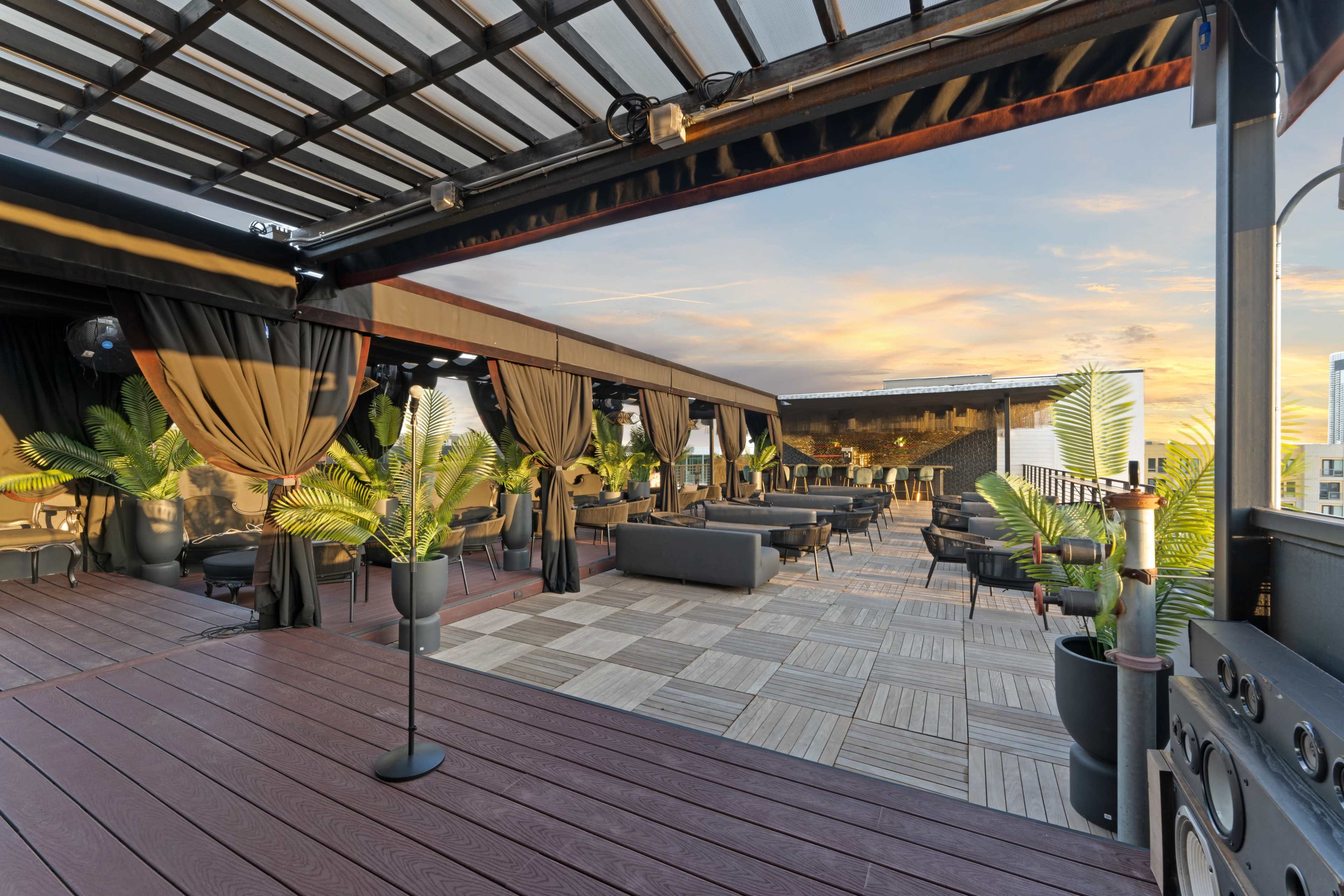 The image depicts a rooftop terrace with seating areas, palm plants, and a sunset sky in the background.