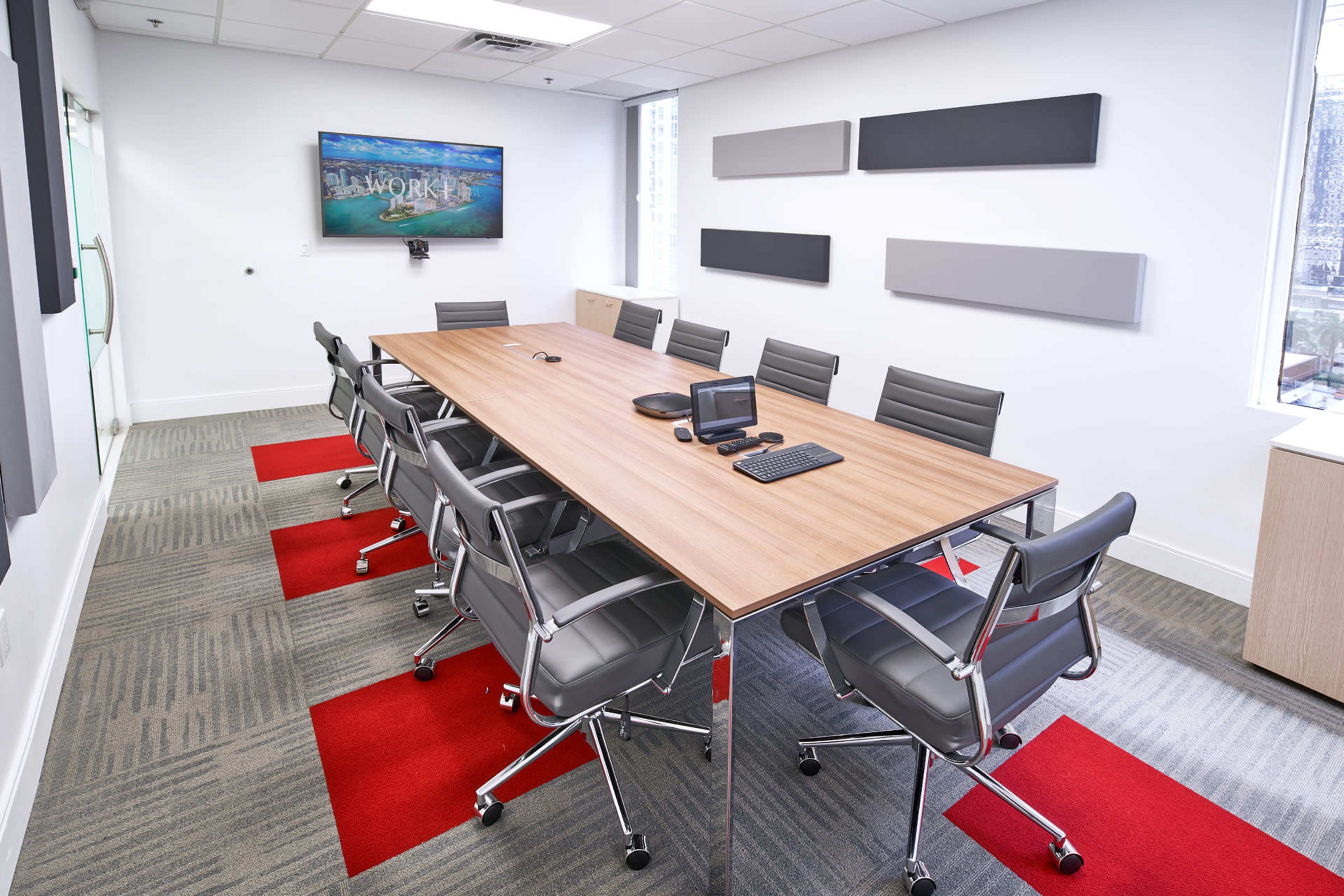 A modern conference room features a large wooden table surrounded by eight gray chairs, with a mounted screen and sound equipment along the wall.