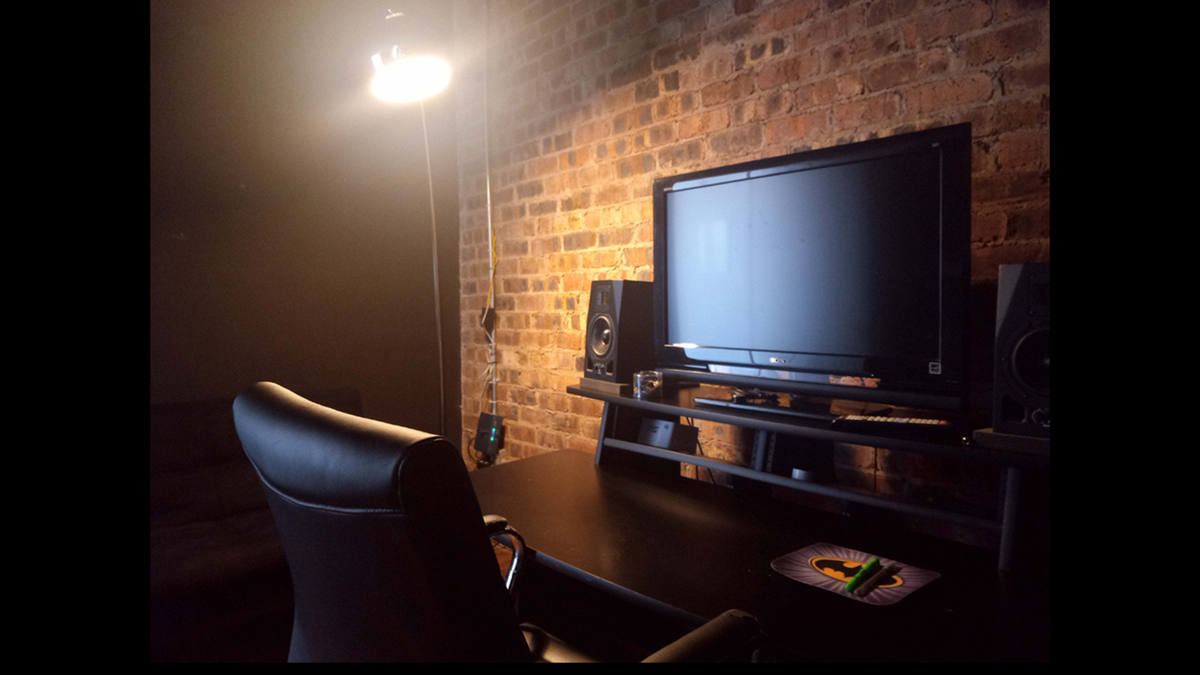 A dark room features a black desk with a computer monitor, speakers, and a swivel chair against a brick wall, illuminated by a hanging light.