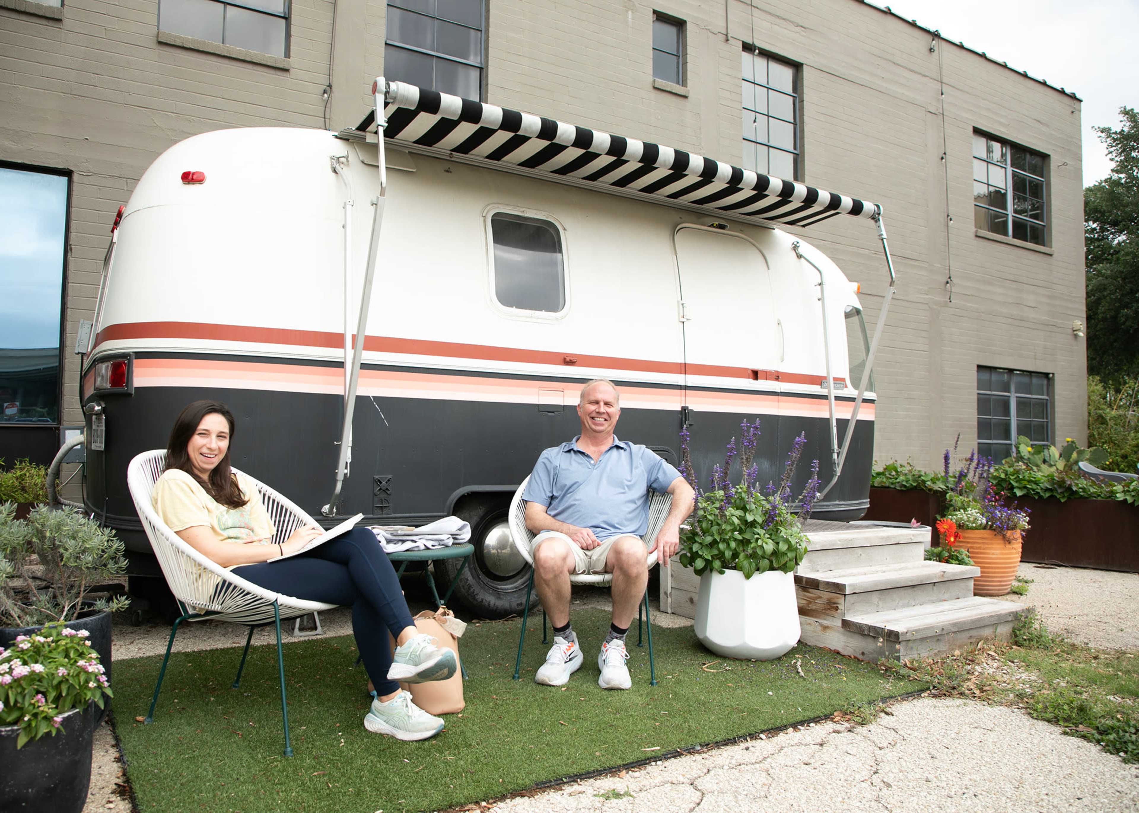 A man and a woman sit outside a vintage trailer, with potted plants and greenery surrounding them.