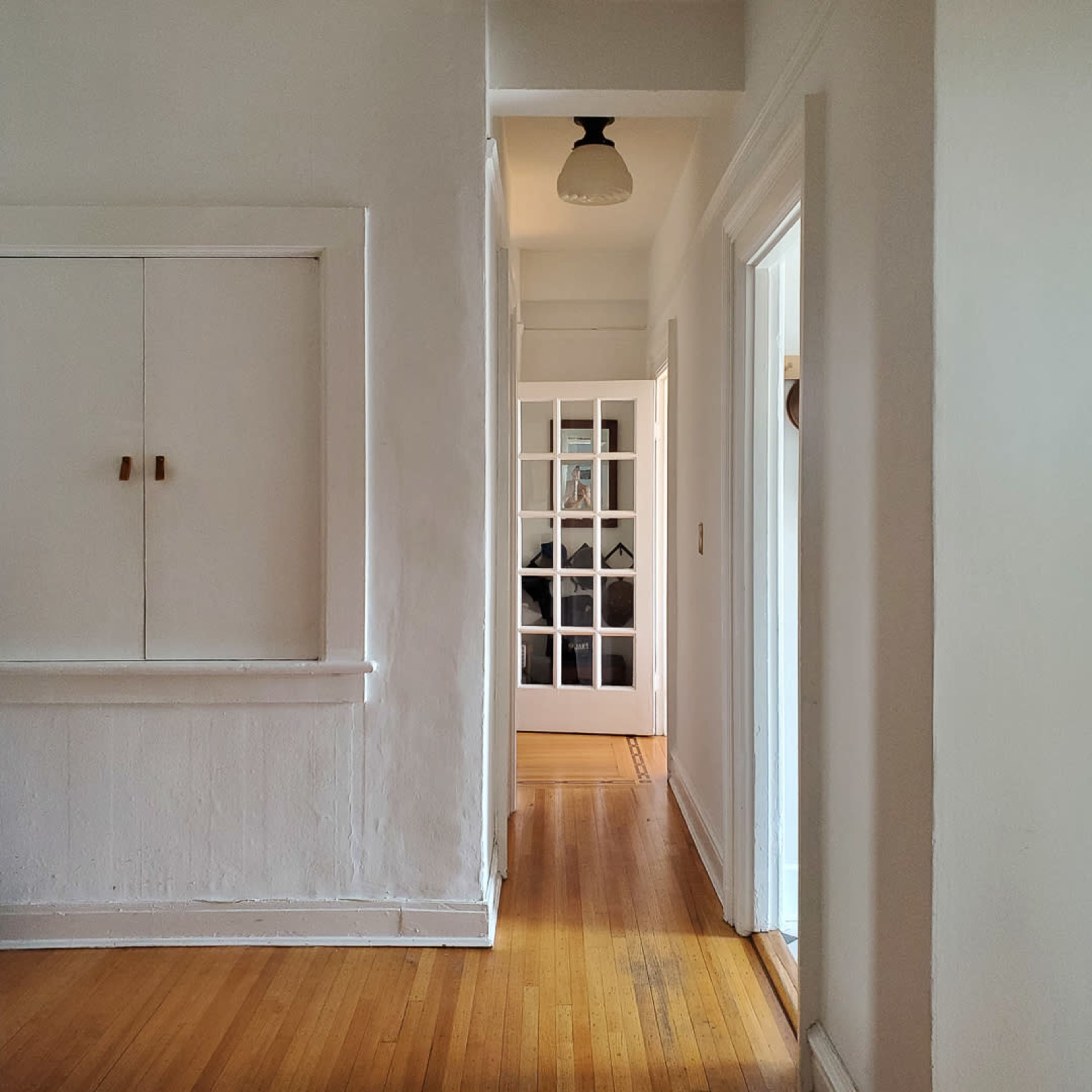 A narrow hallway with white walls, wooden floors, and a door at the end featuring glass panels.