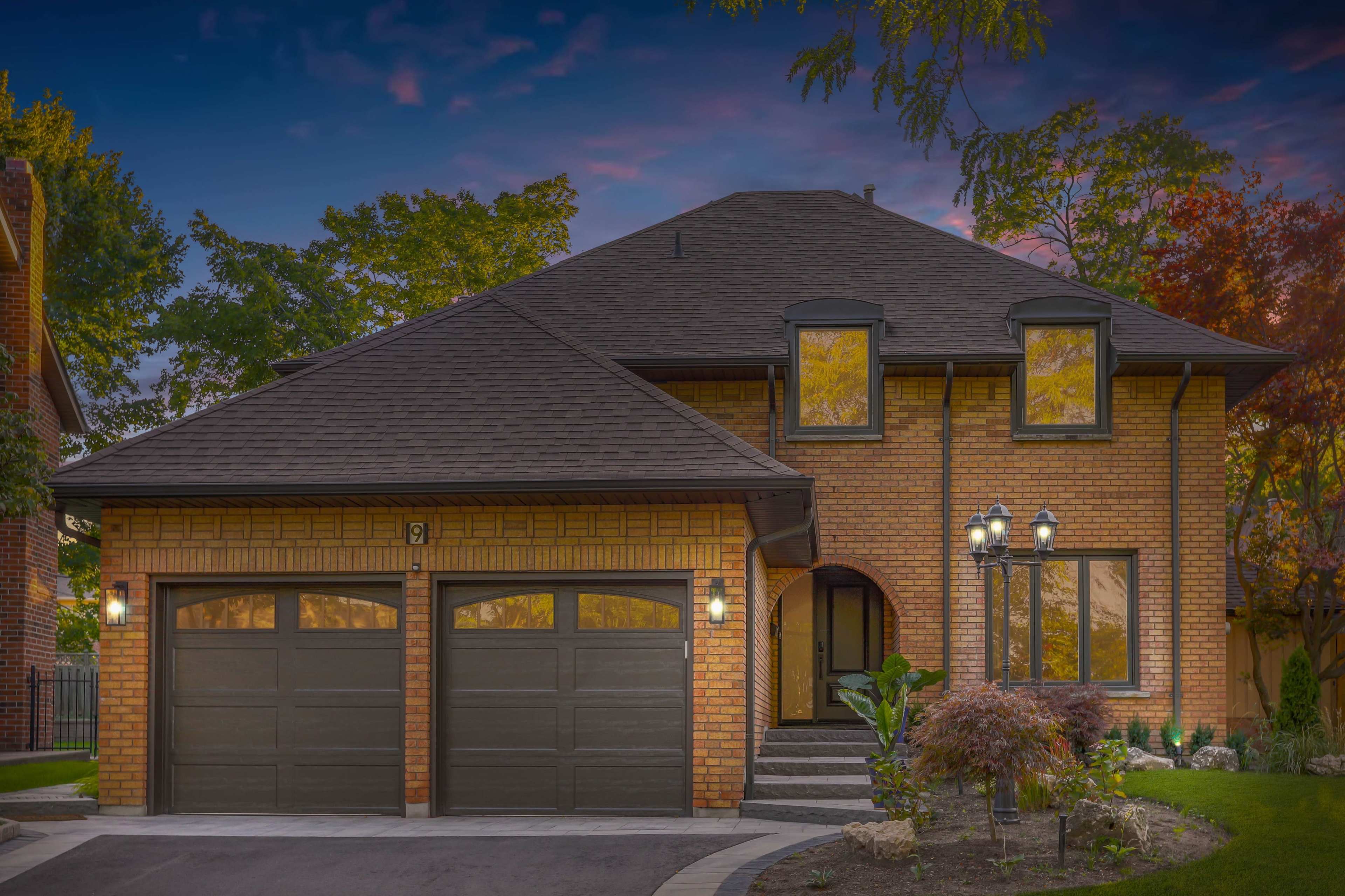 A large brick house with a steep roof, two garage doors, and landscaped greenery in front.