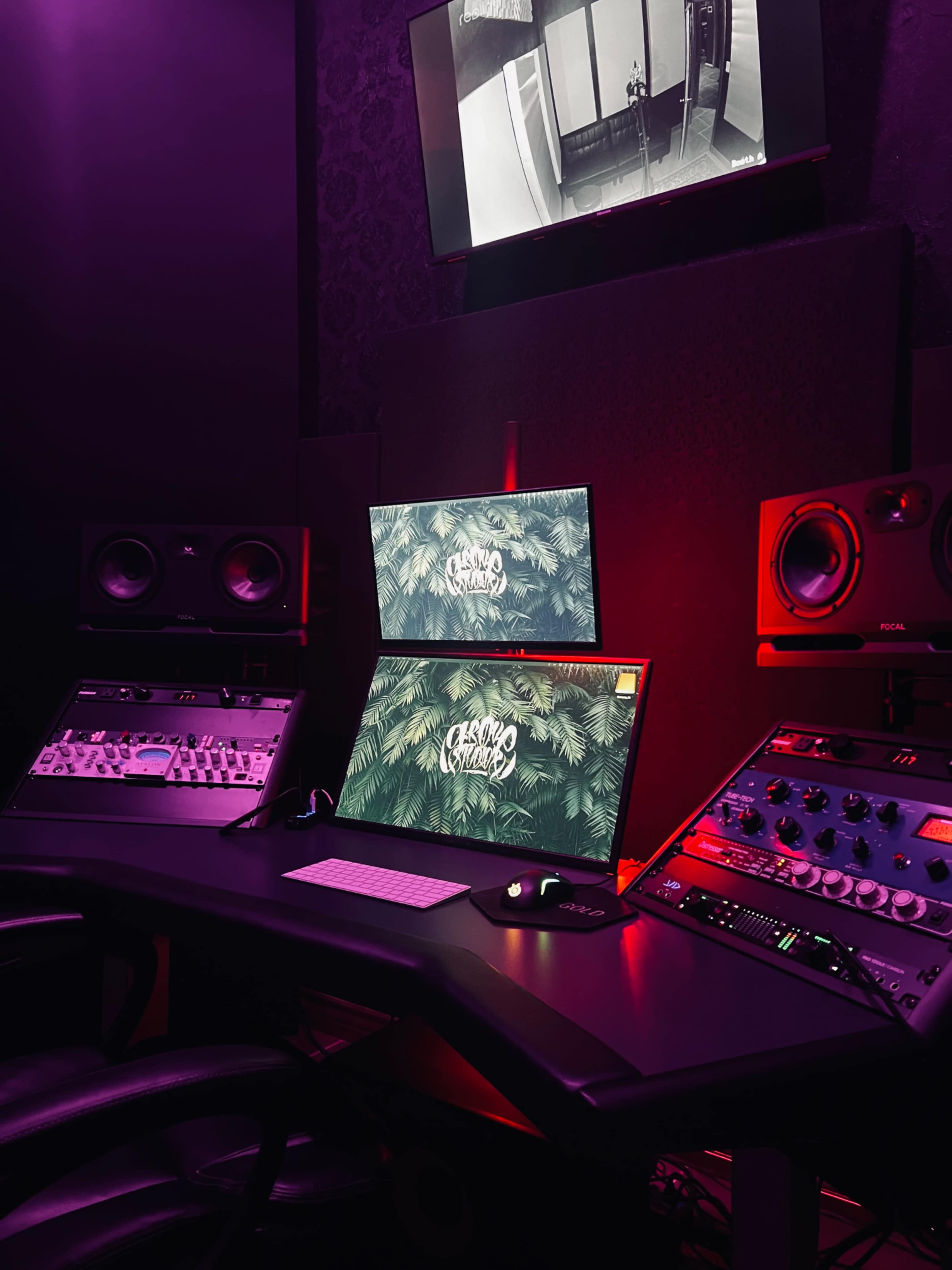 The image shows a dimly lit music production studio featuring two computer monitors, audio equipment, and a black desk with a keyboard and mouse.