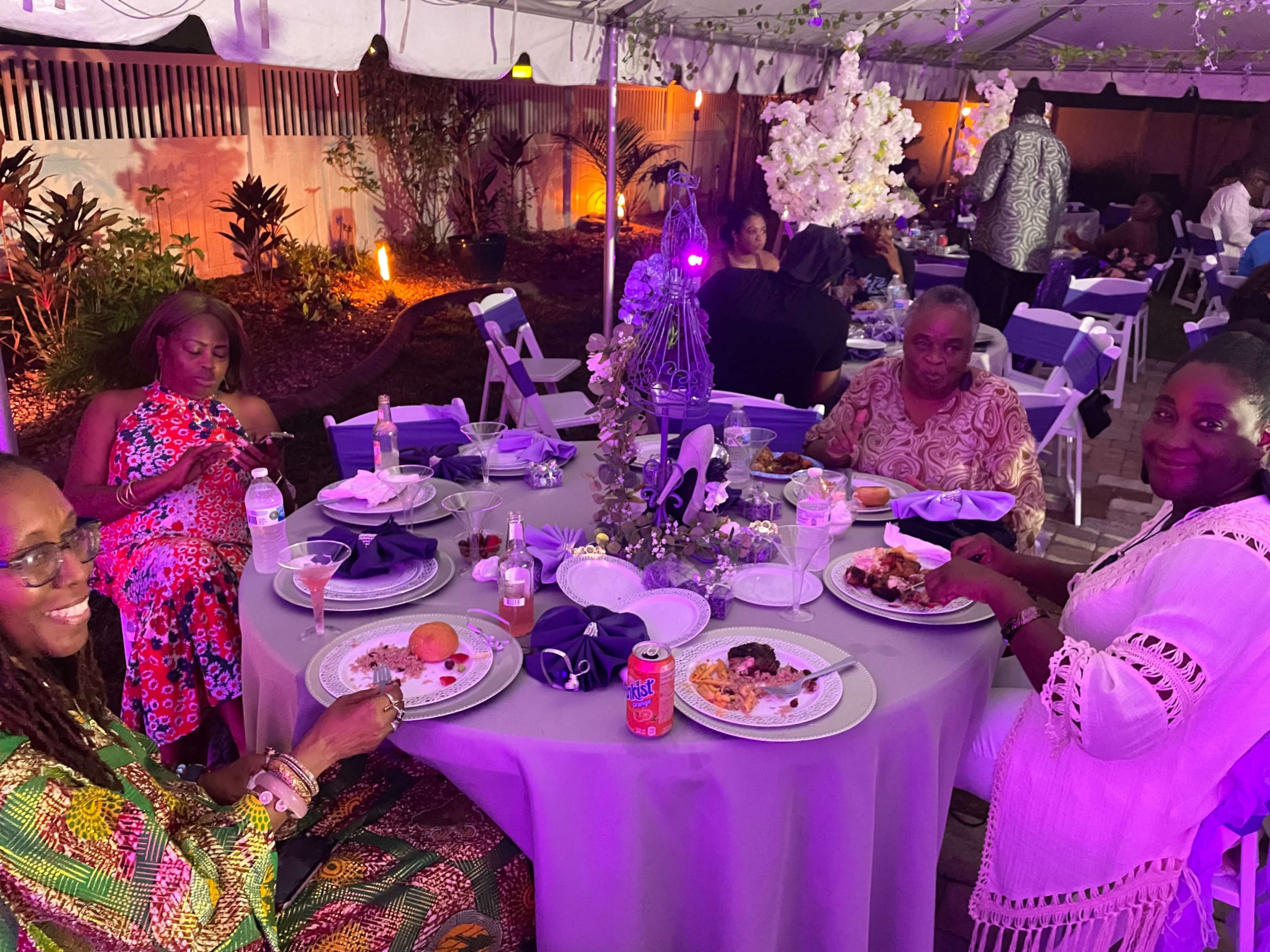 A group of four women sits around a table adorned with food and drinks at an outdoor dining event under a tent.