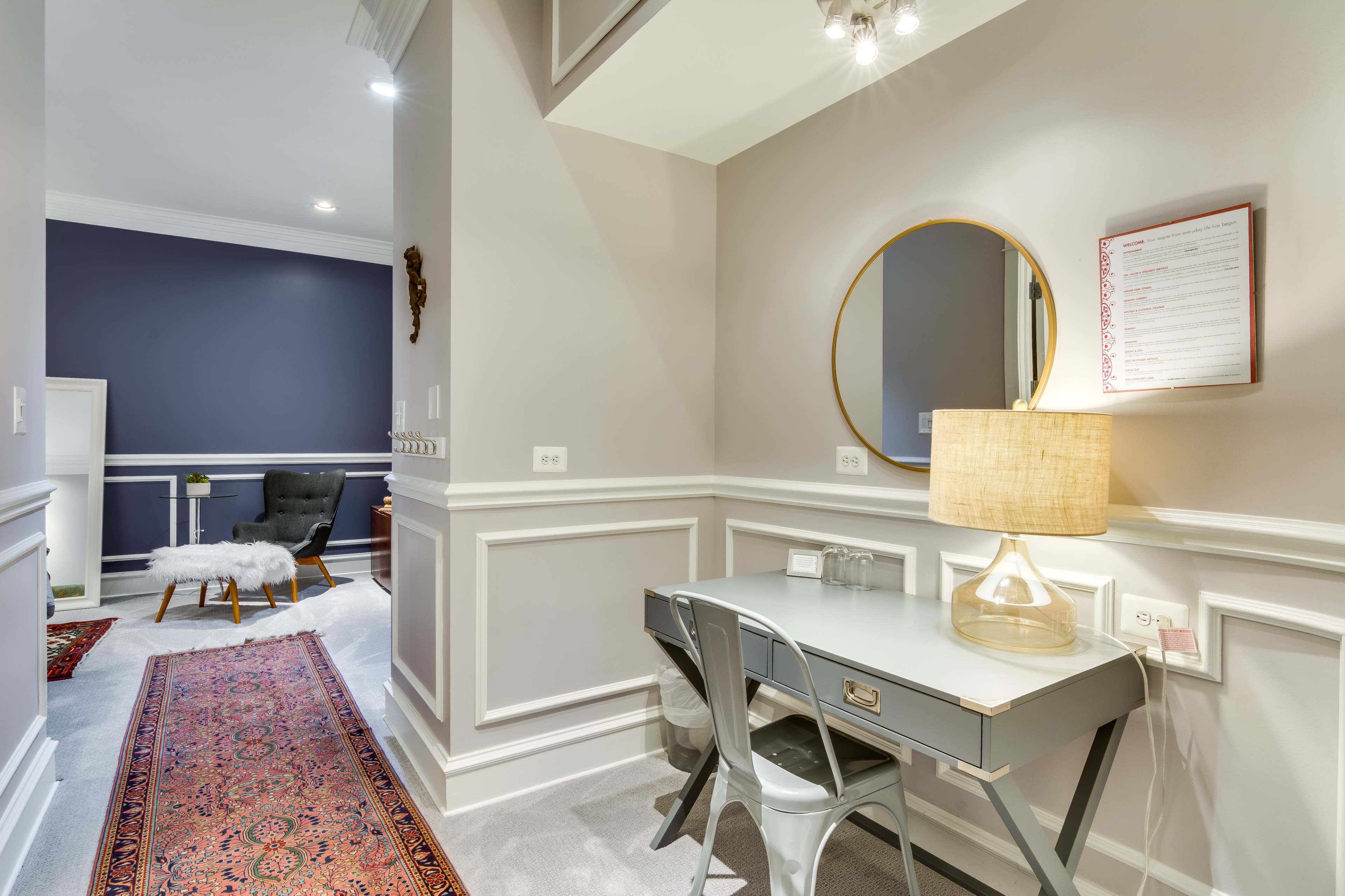 A well-lit hallway with a small desk, a round mirror, a lamp, and decorative elements including a rug and a chair in the background.