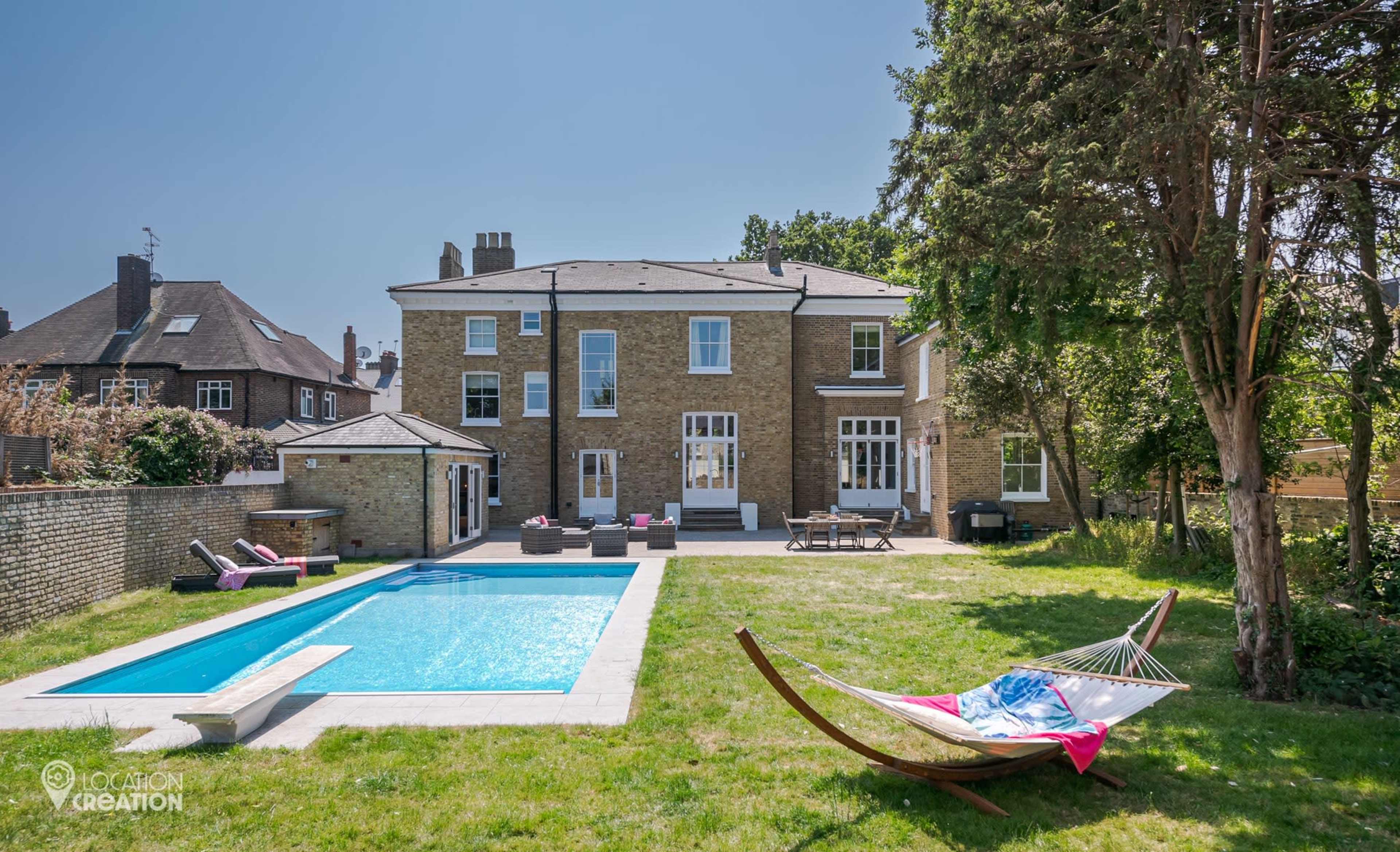A large stone house with a swimming pool and a hammock is set in a grassy backyard.