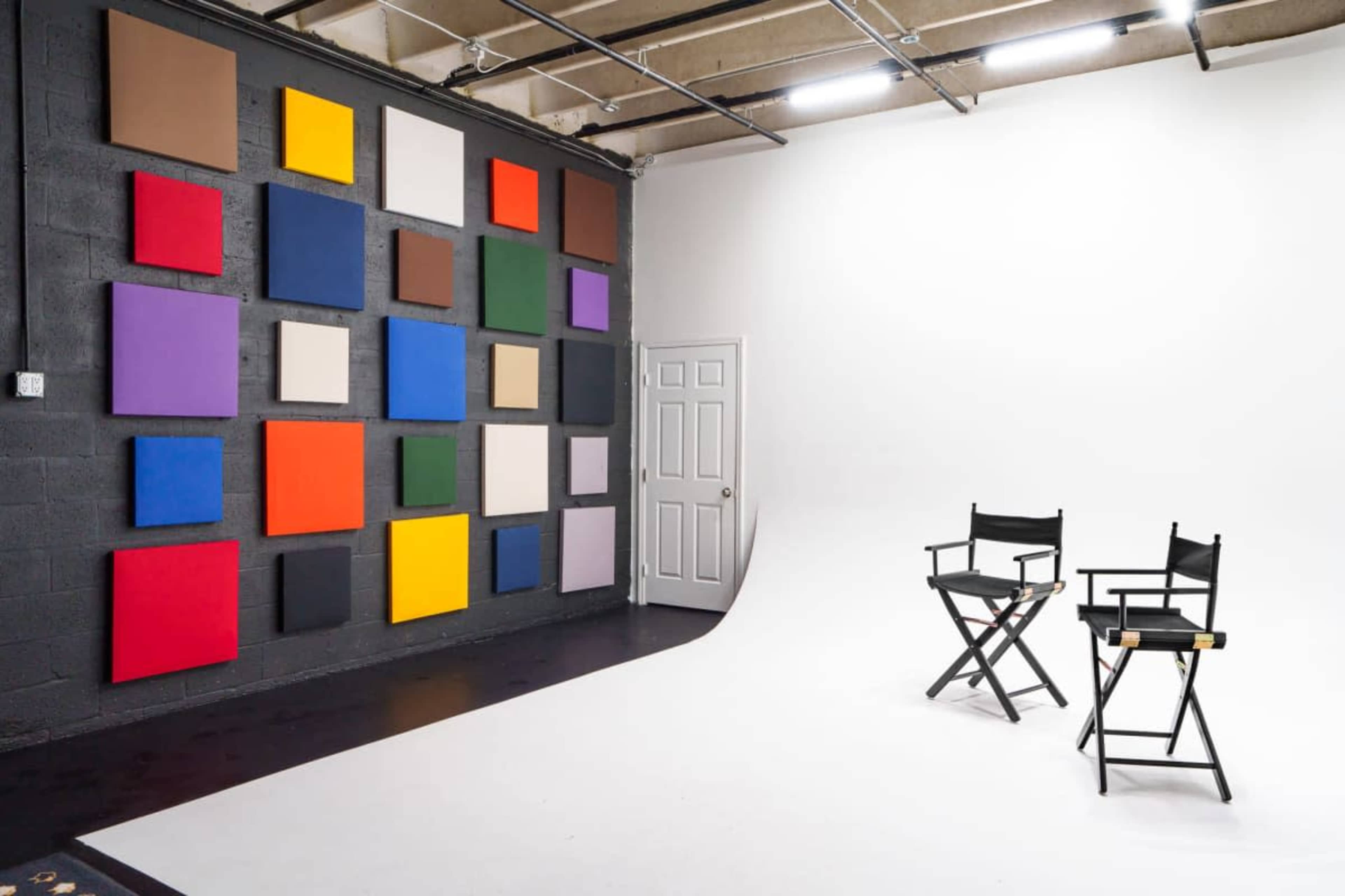 The image shows a photography studio with a white seamless backdrop and two director's chairs in front of a wall featuring colorful canvases arranged in a grid pattern.