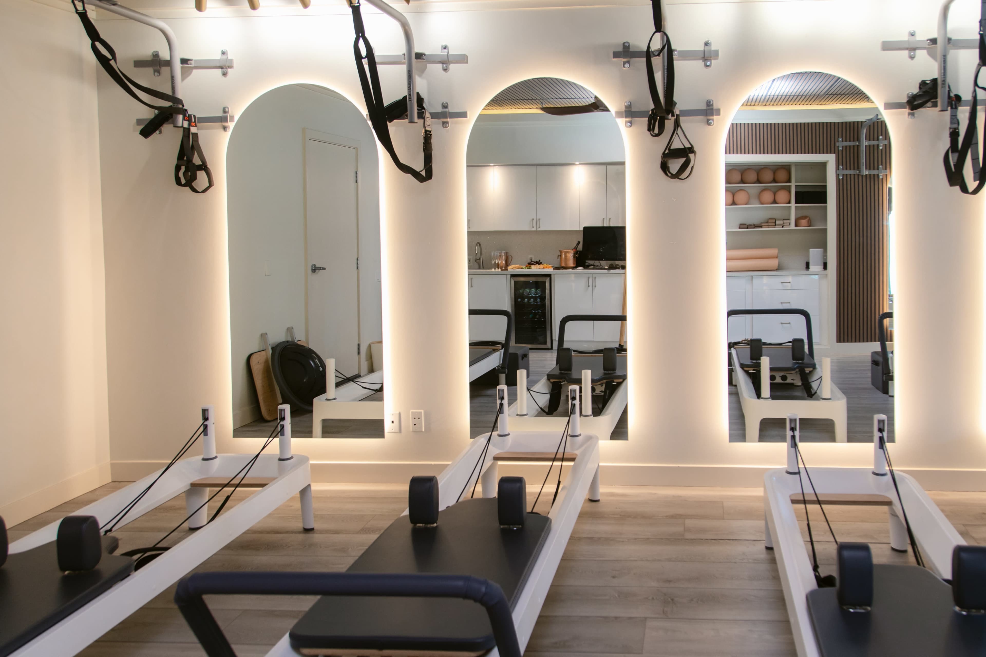 The image shows a fitness studio with sleek reformer Pilates machines arranged in front of illuminated mirrors and a modern kitchen area visible in the background.