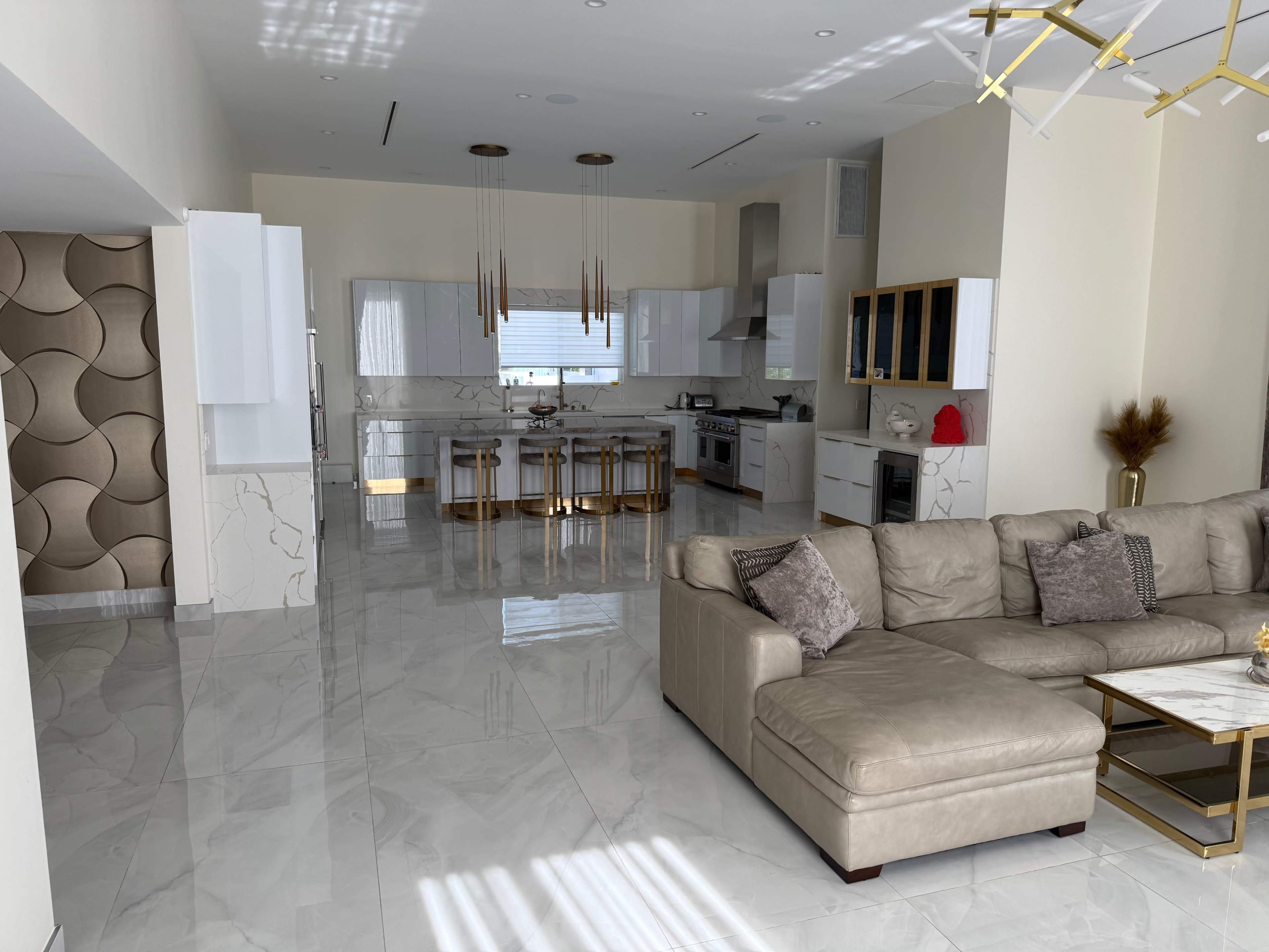 The image shows a spacious, modern kitchen and living area with marble floors, sleek cabinetry, and a large sectional sofa.