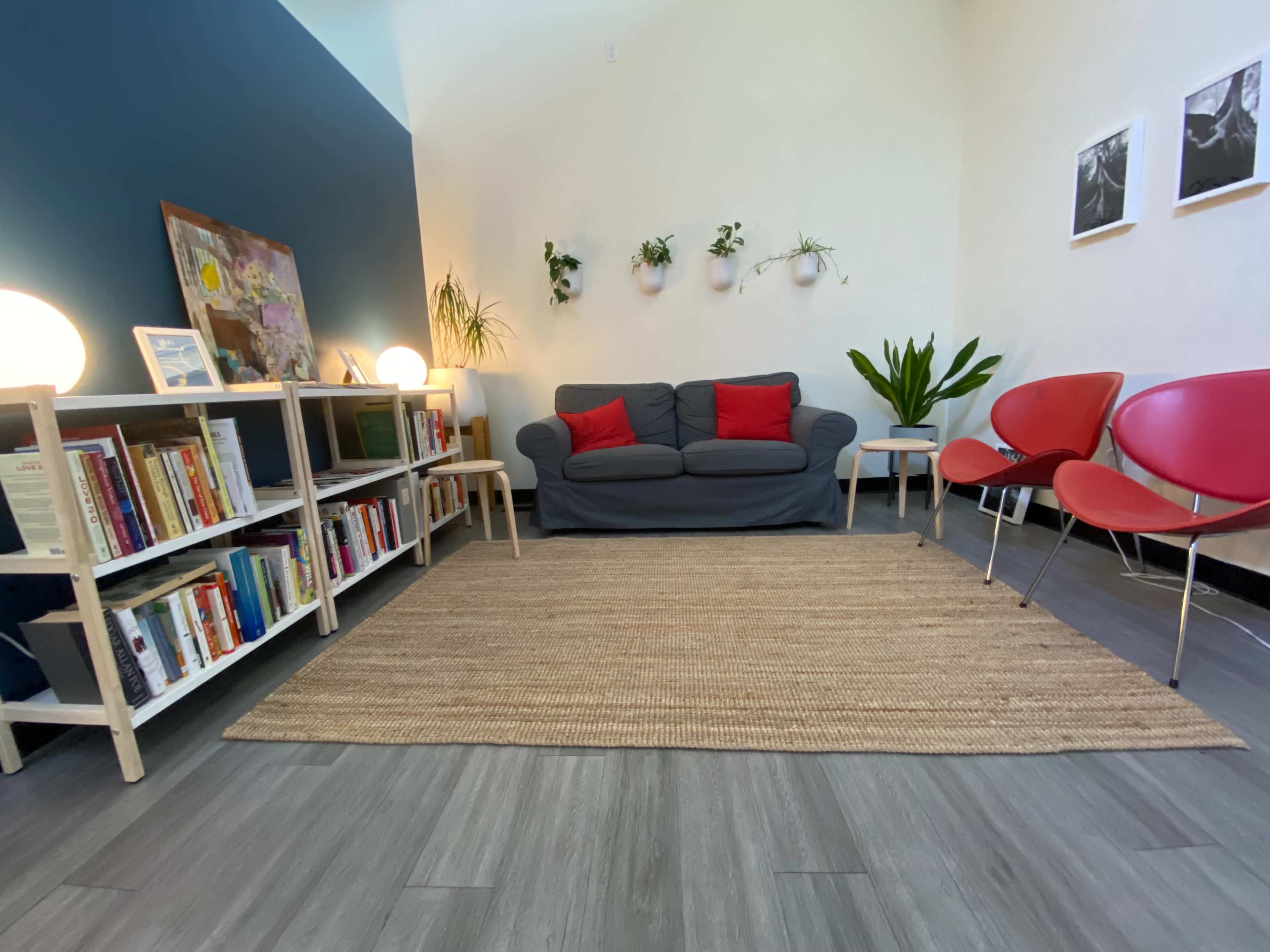 The image shows a cozy reading nook with a gray sofa, red pillows, a rug, shelves filled with books, and red chairs against a blue and beige wall with plants hanging.