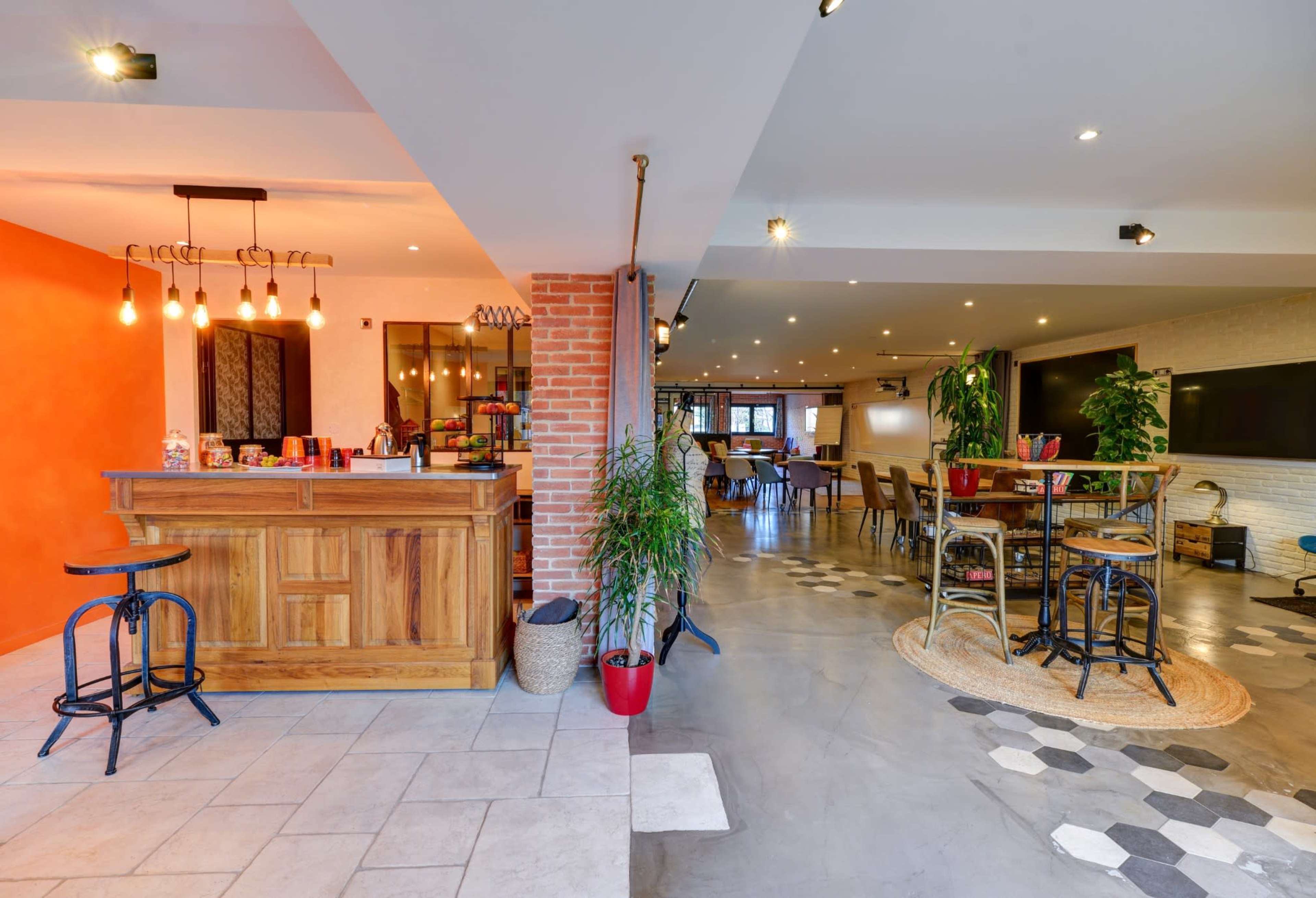 A modern interior space featuring a wooden bar area with a vibrant orange wall, leading into a dining area with varied seating arrangements and decorative plants.
