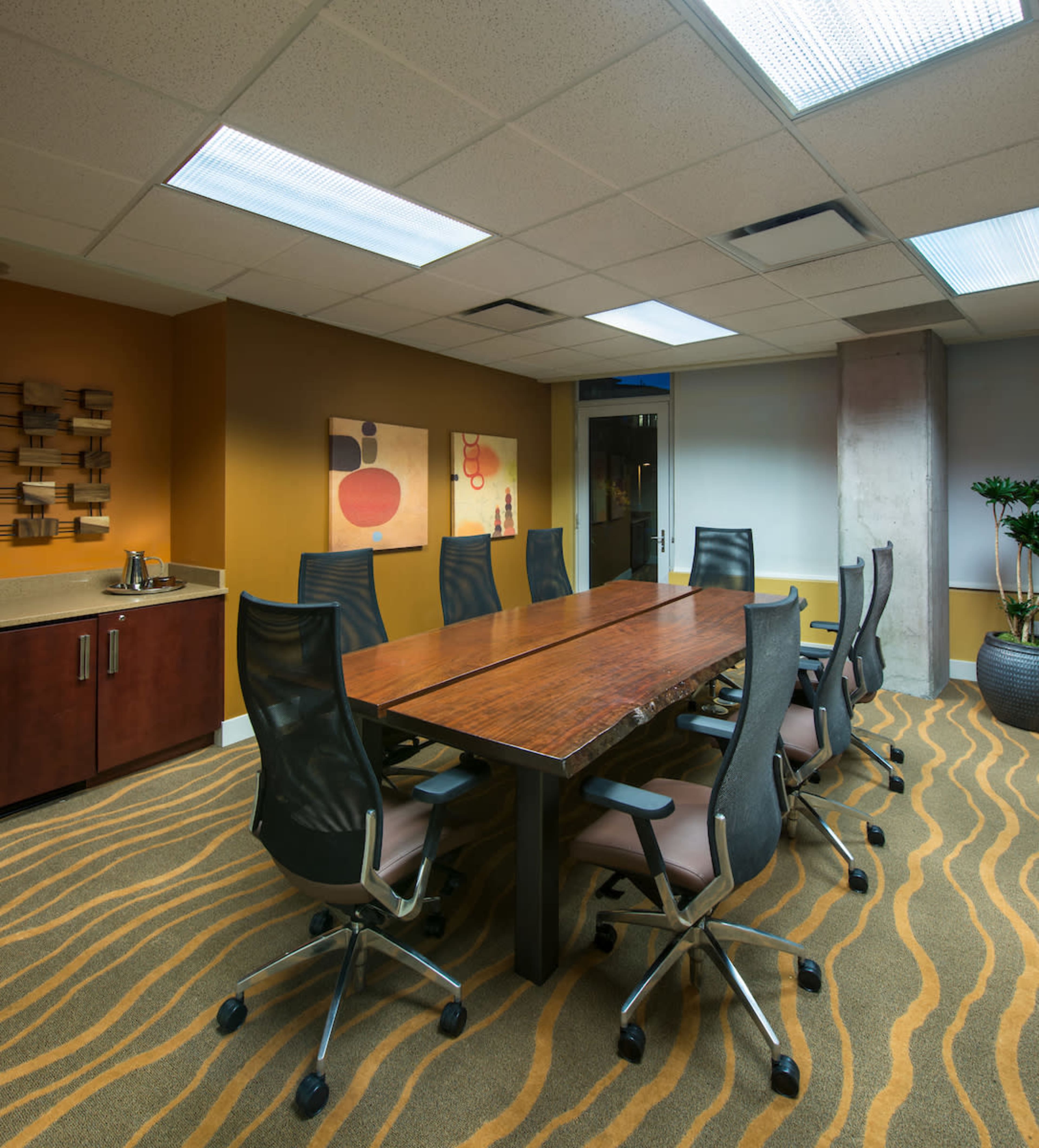 The image shows a modern conference room featuring a large wooden table surrounded by ergonomic chairs, with abstract art on the walls and a carpet with a wavy pattern.