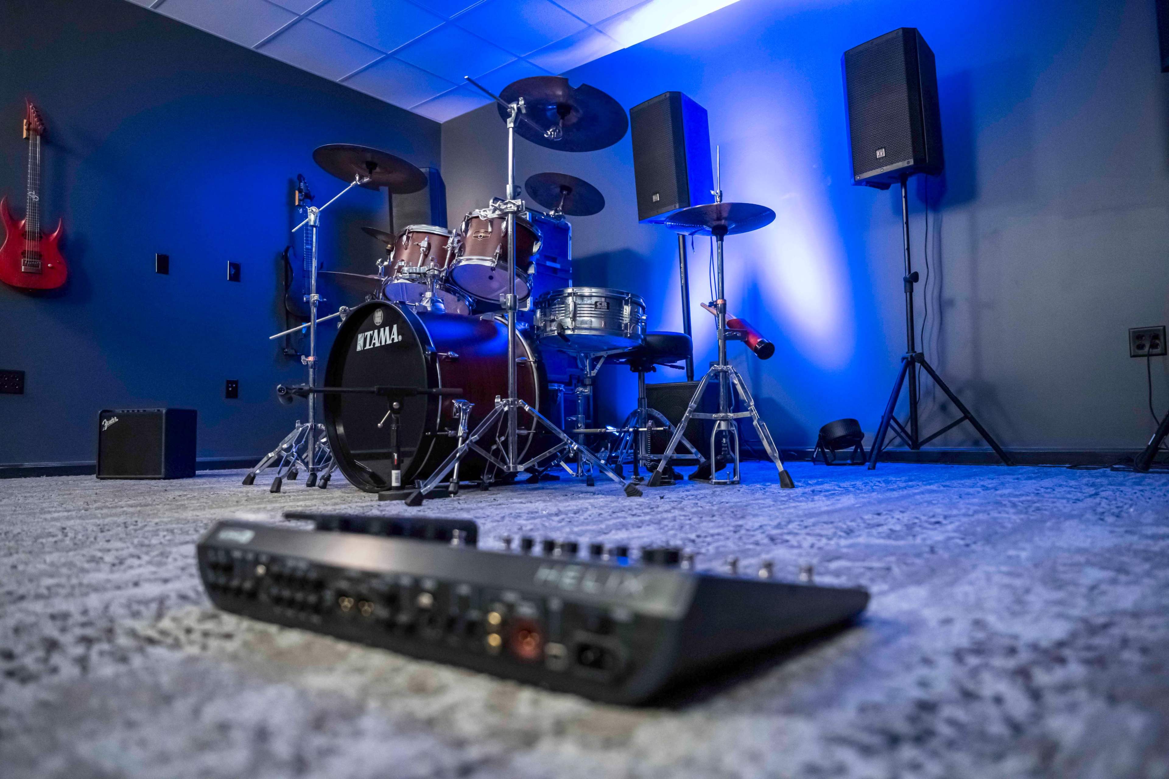 A music studio with a drum set, speakers on stands, and an audio mixer placed on a carpeted floor.