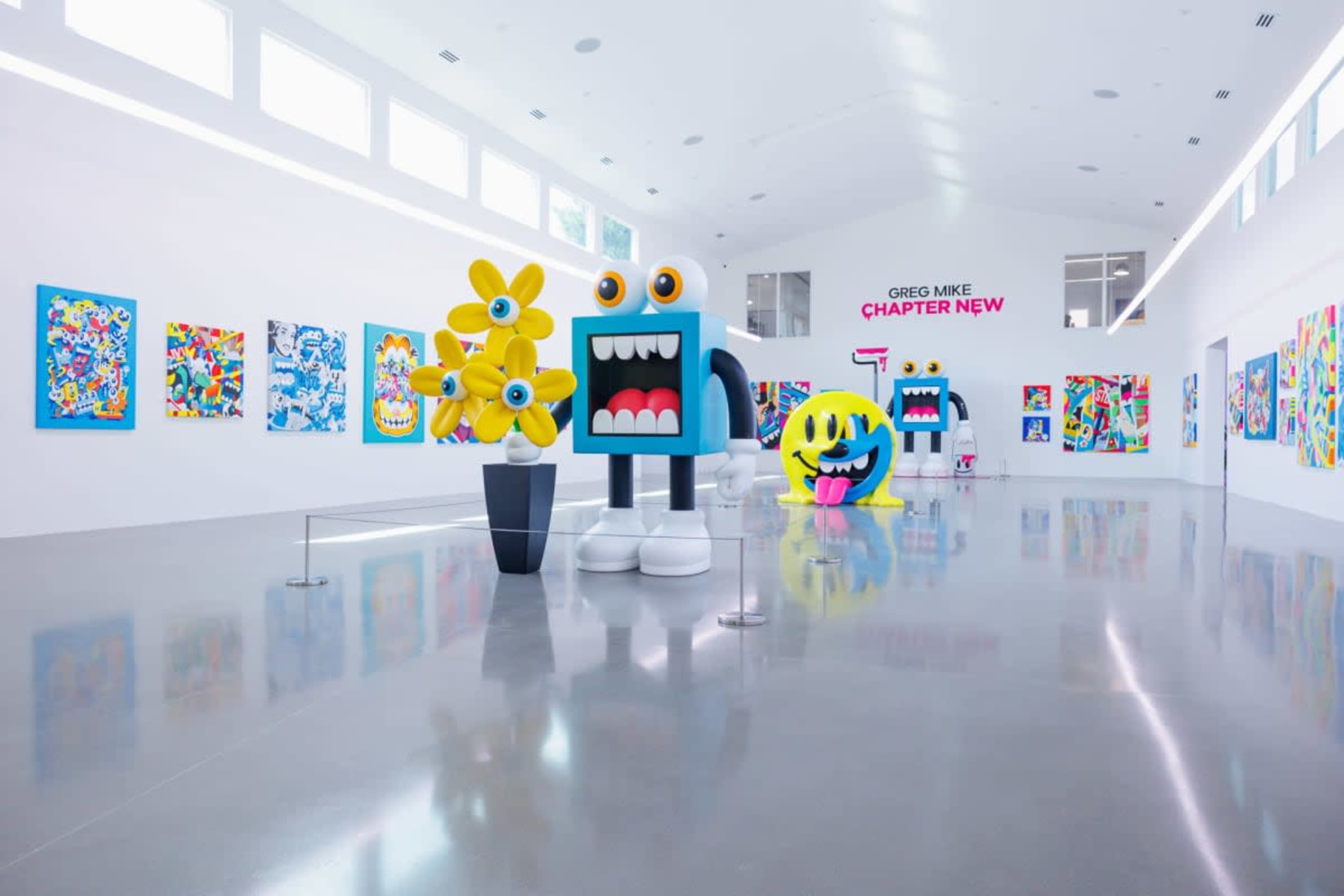 The image shows a brightly lit gallery featuring vibrant artwork on the walls, with large, colorful sculptures on display in the center.