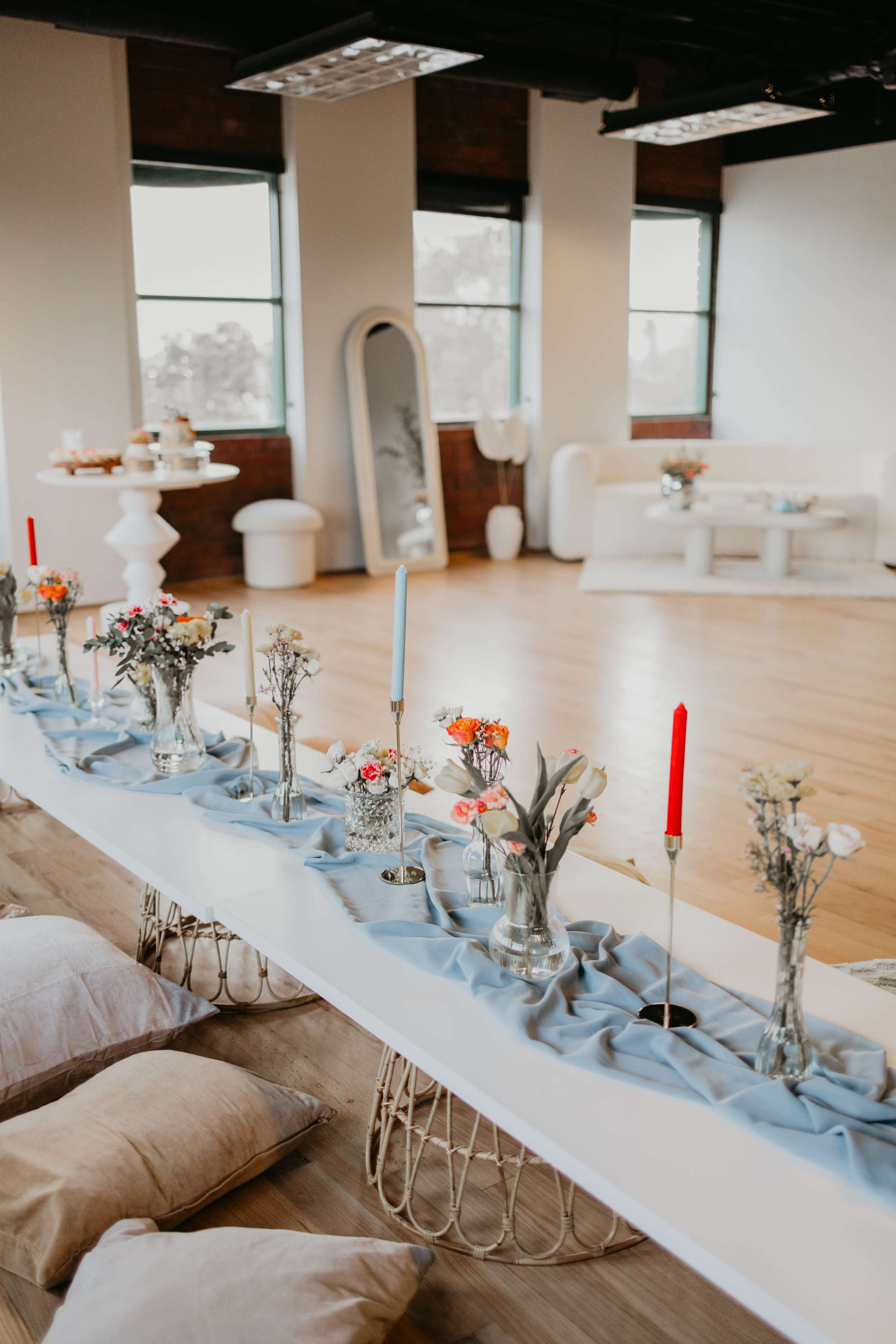 The image shows a spacious, modern event space featuring a long table with a light blue runner, adorned with various vases of flowers and candles, alongside decorative pillows on the floor.