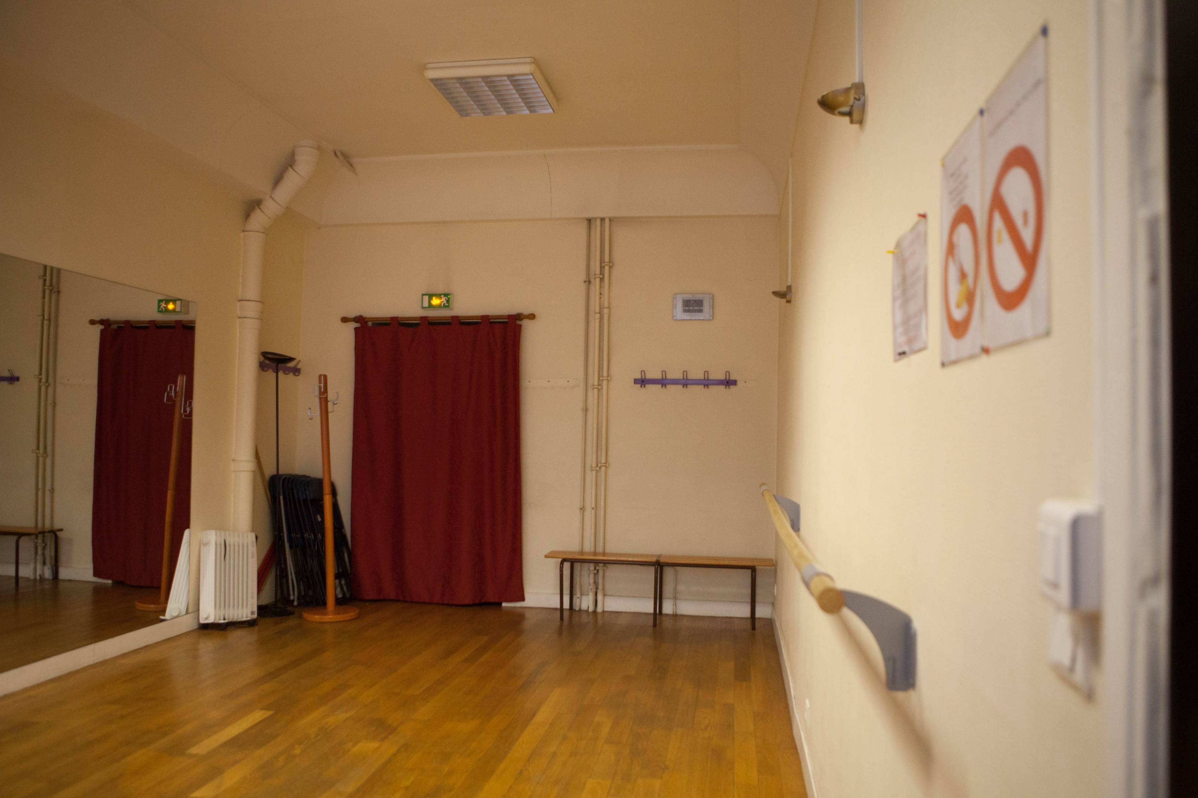 The image shows a dance studio with wooden flooring, a wall-mounted ballet barre, red curtains, and signs indicating rules.