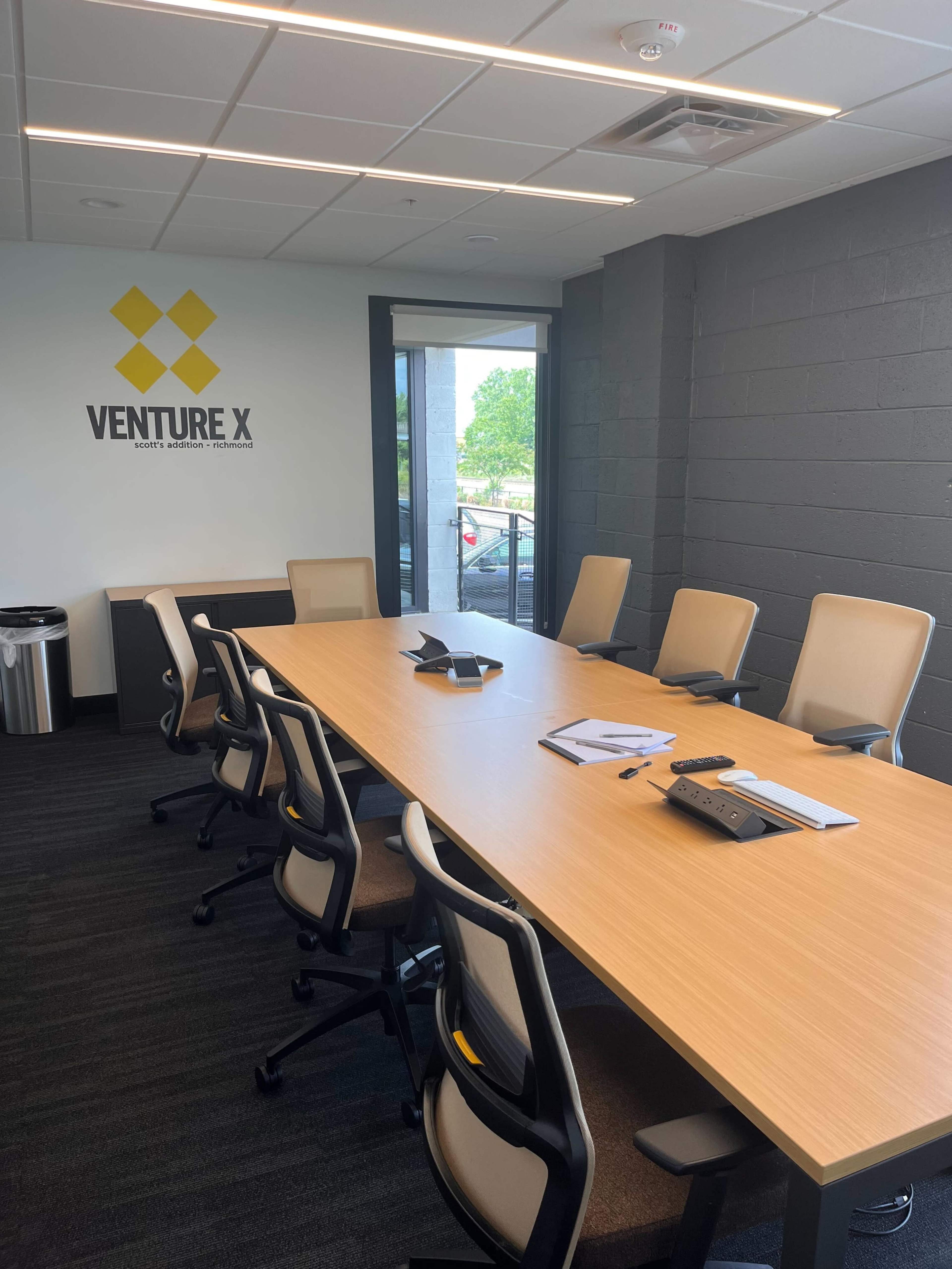 A spacious conference room features a long wooden table surrounded by ergonomic chairs, with a wall displaying the "Venture X" logo.