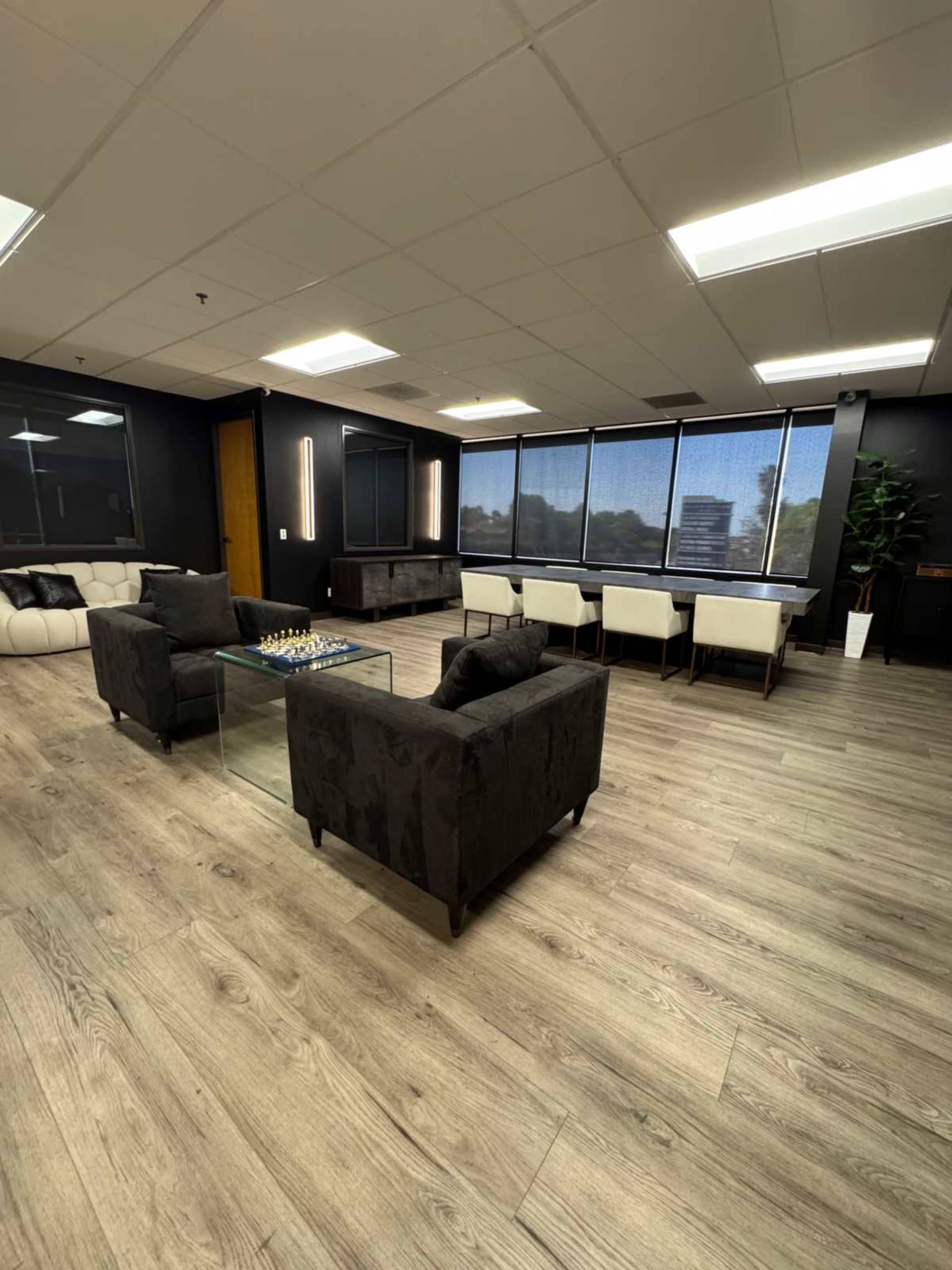 The image shows a modern office space featuring a seating area with black sofas, a glass coffee table, and a dining area with white chairs near large windows.
