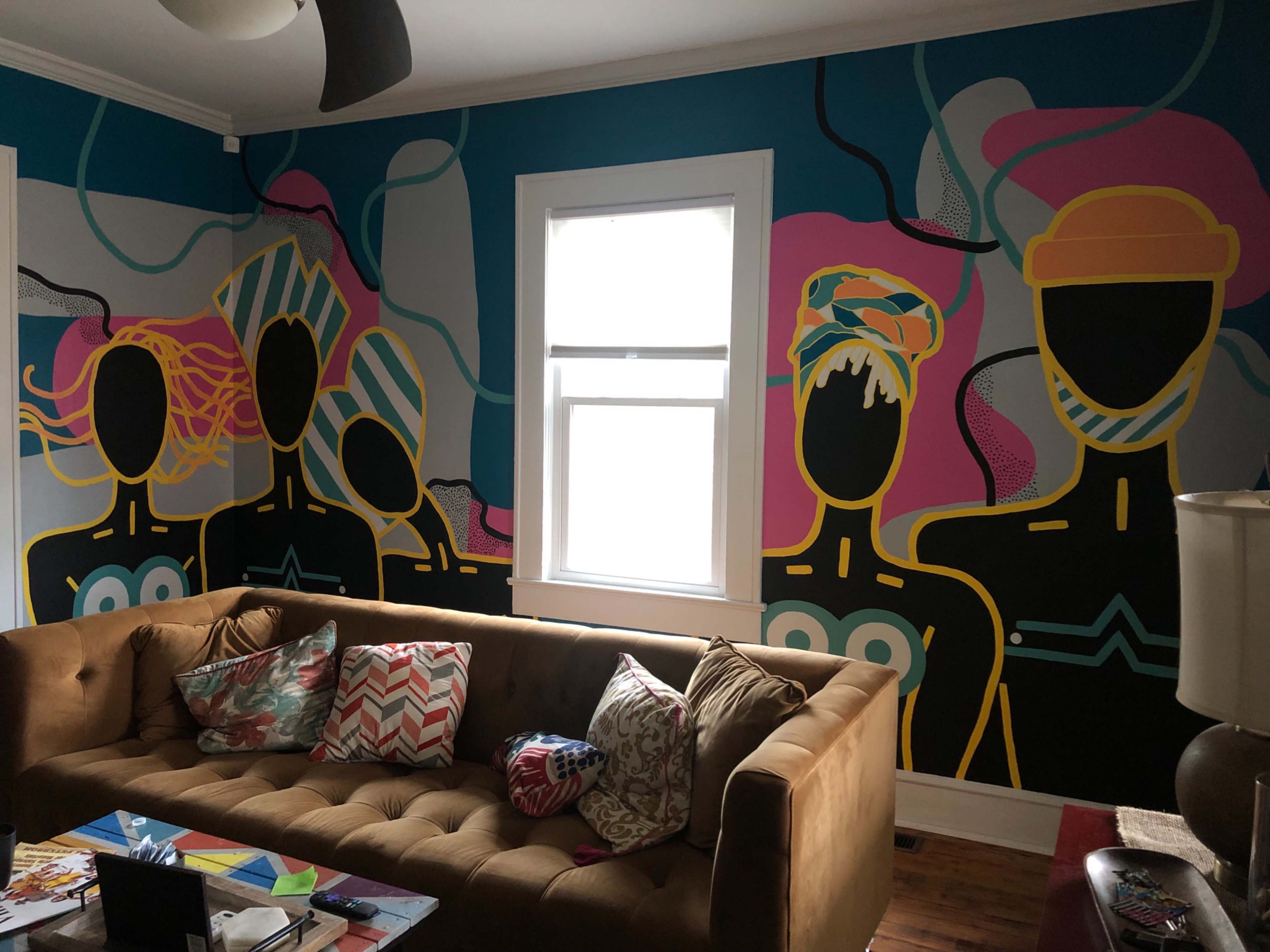 A living room with a brown sofa, colorful pillows, and a wall mural depicting three abstract figures with geometric patterns.