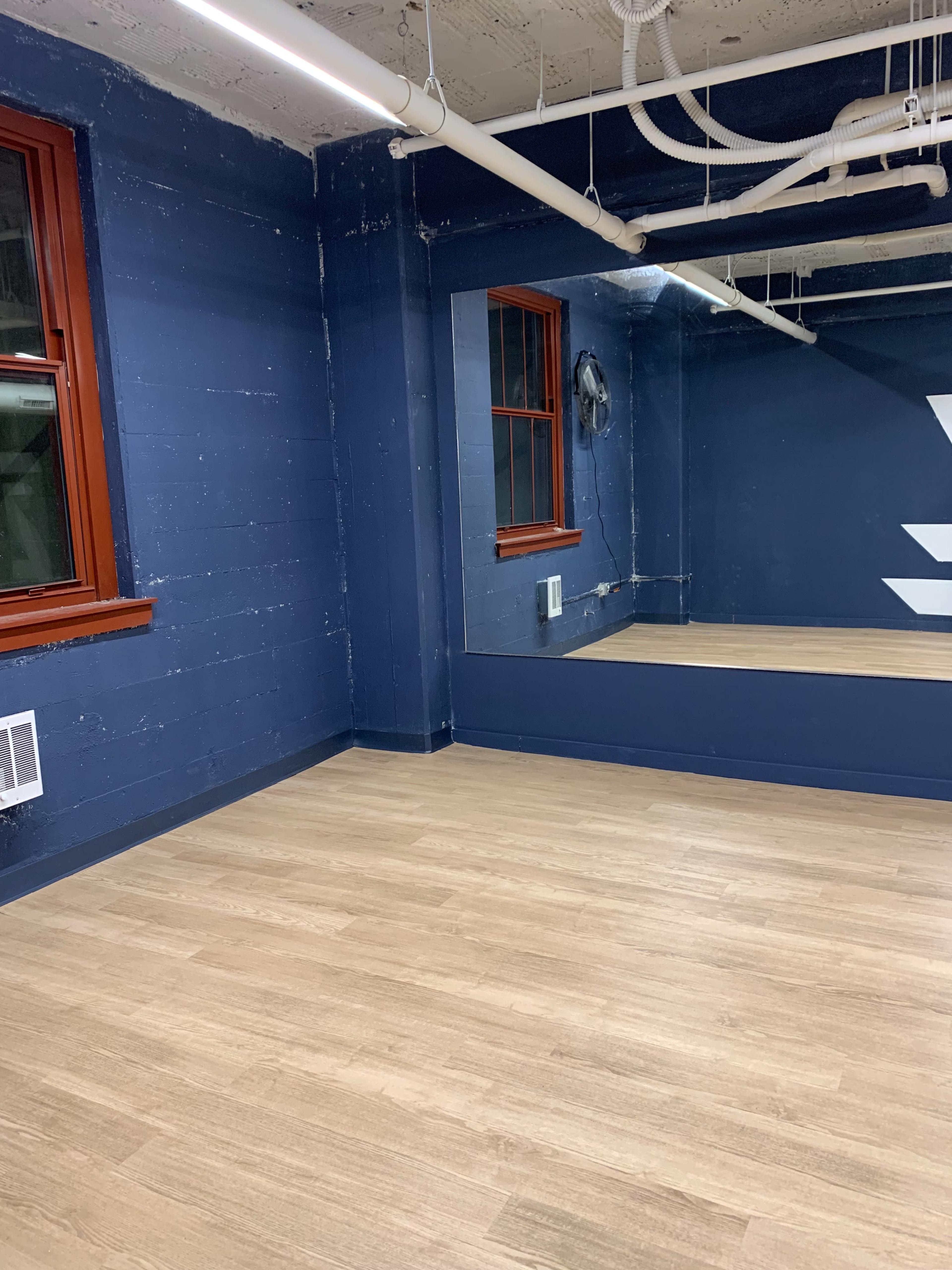 A room with blue walls, wooden flooring, a large mirror, and two windows with red frames.