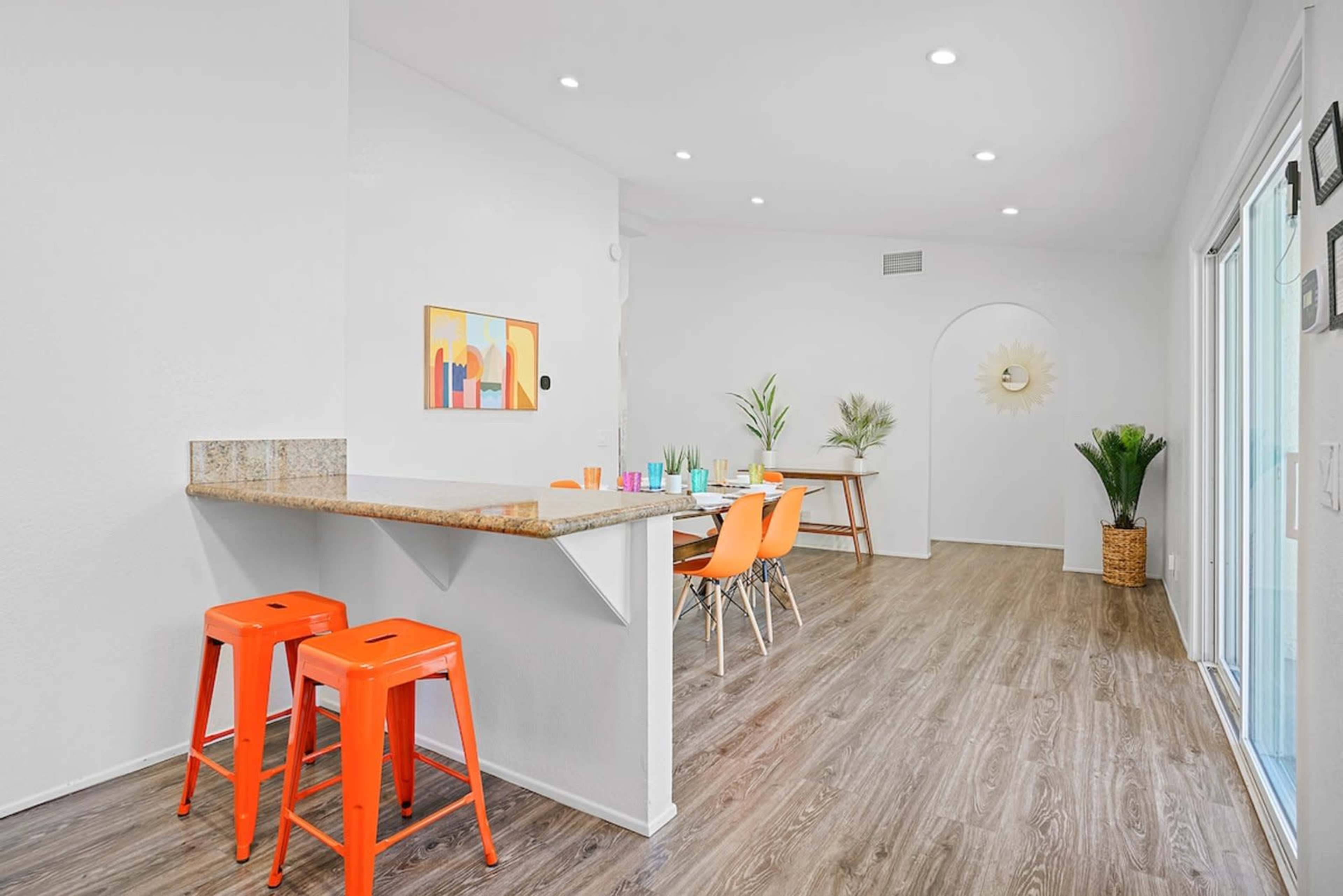 The image shows a bright, modern kitchen and dining area with orange bar stools, a dining table with colorful chairs, and light wood flooring.