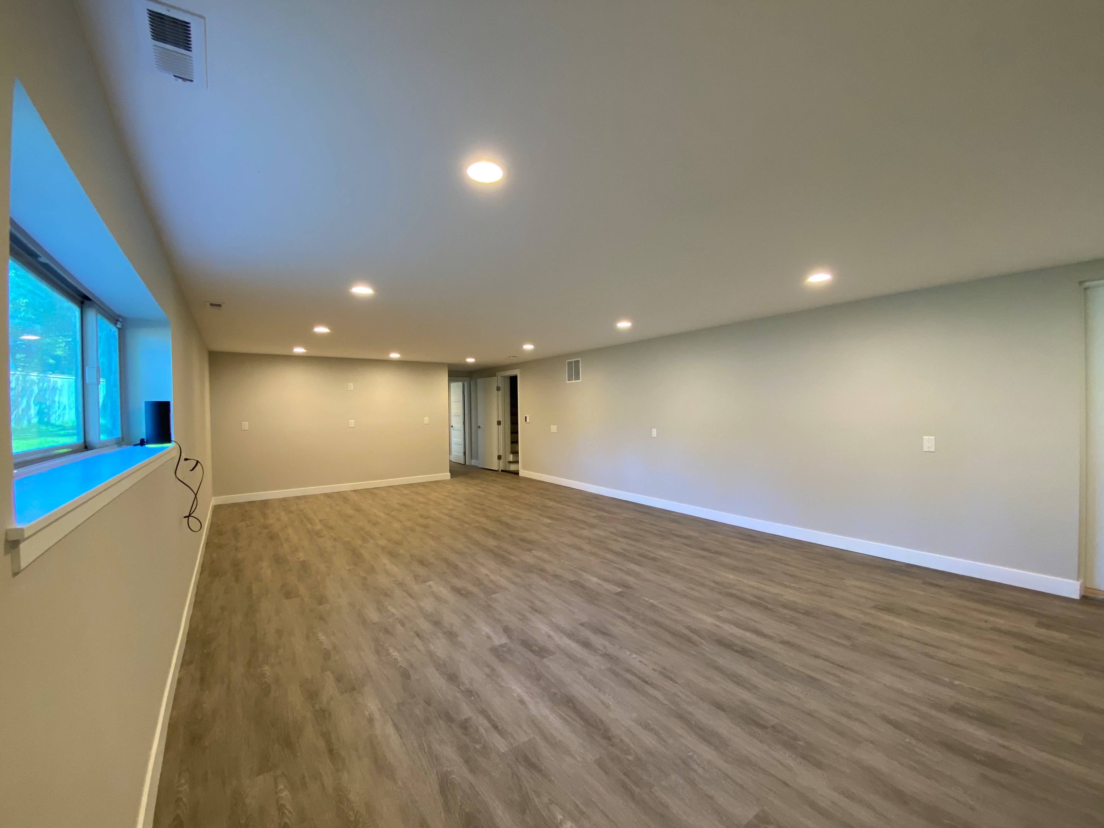 The image shows a spacious, empty room with light-colored walls, a wooden floor, and a large window providing natural light.