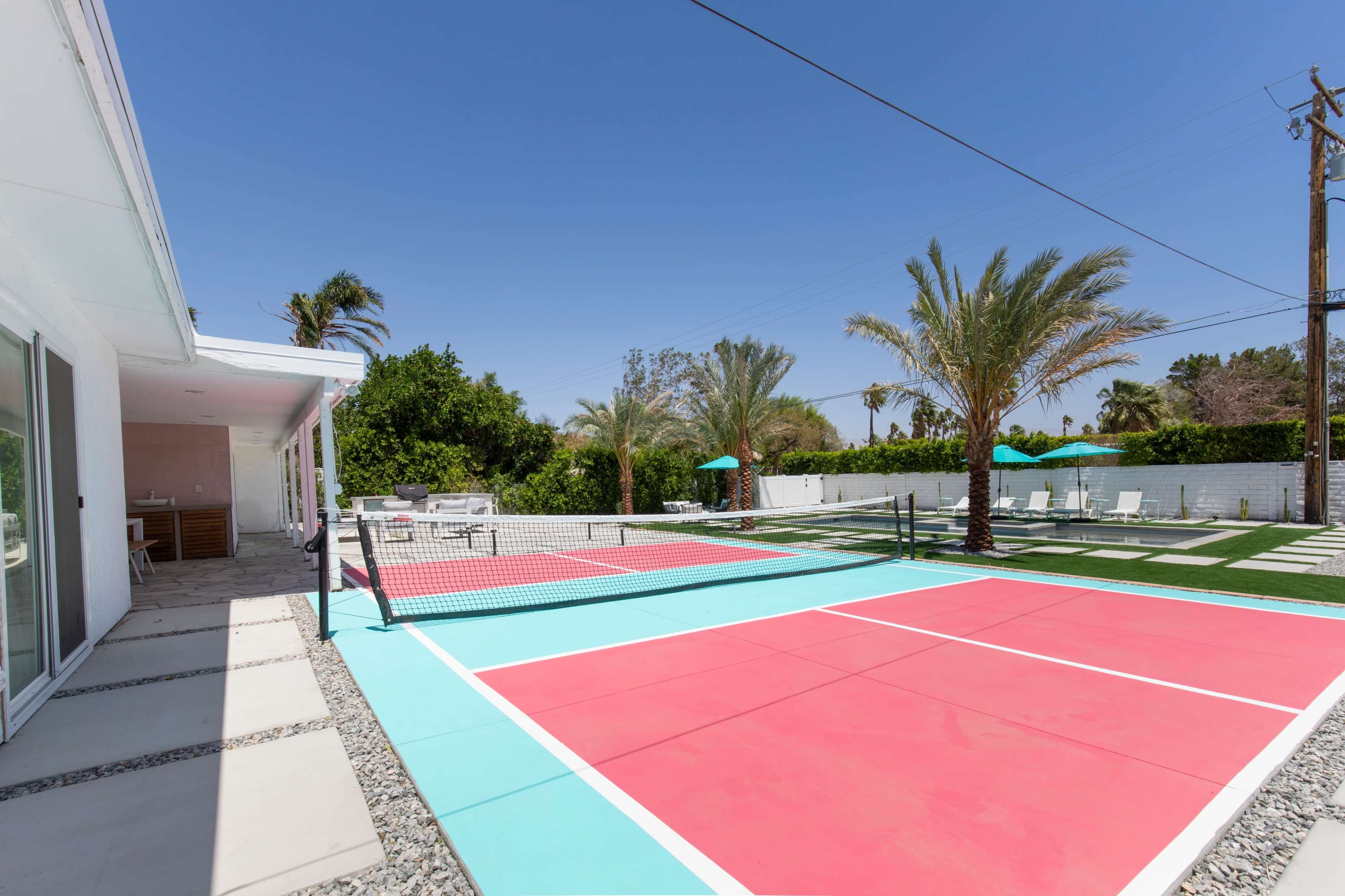 The image shows a brightly colored tennis court in a sunny backyard, surrounded by palm trees and outdoor seating areas.