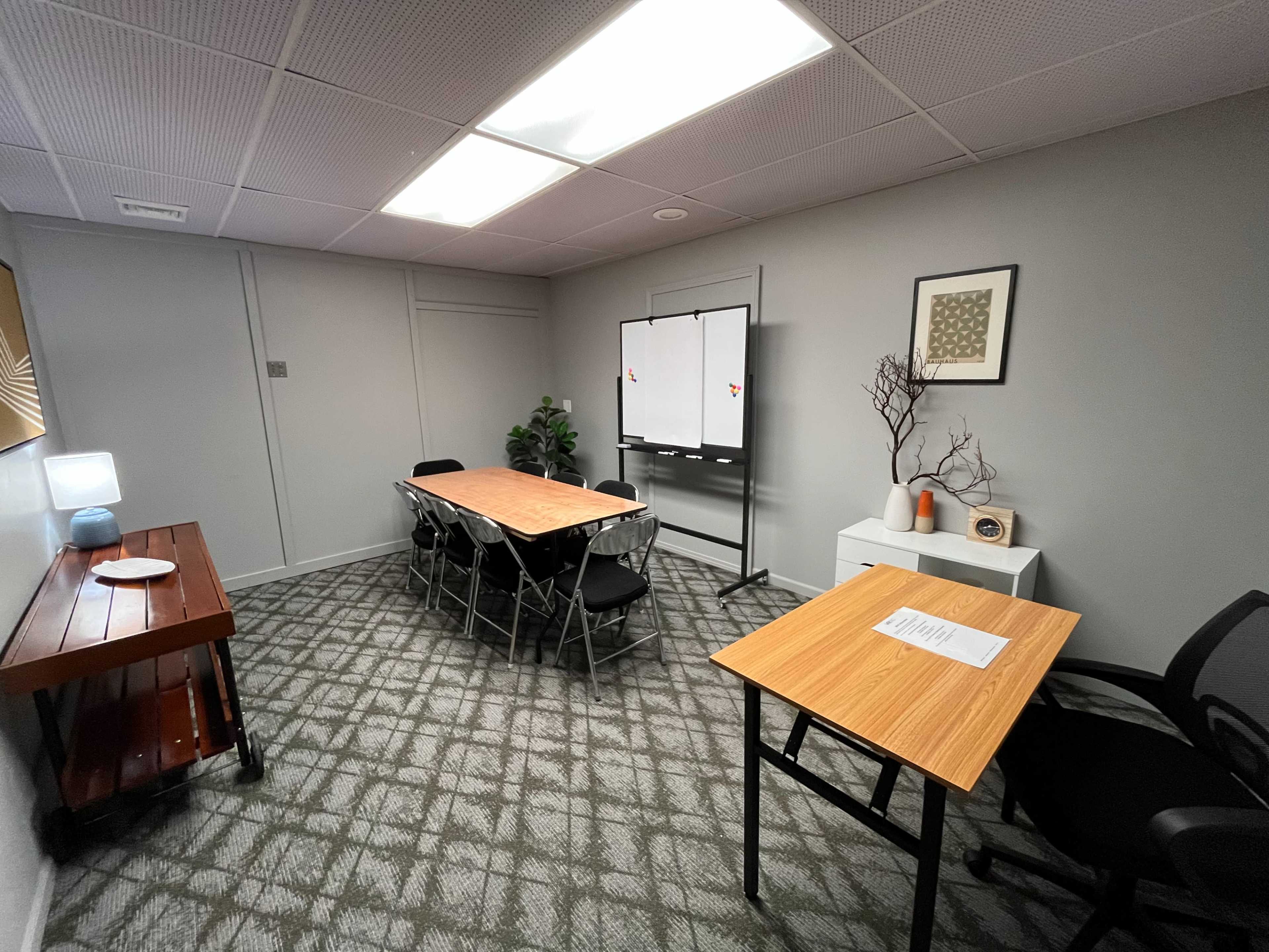 The image shows a small conference room with two tables, several chairs, a whiteboard on a stand, and a decorative plant in the corner.