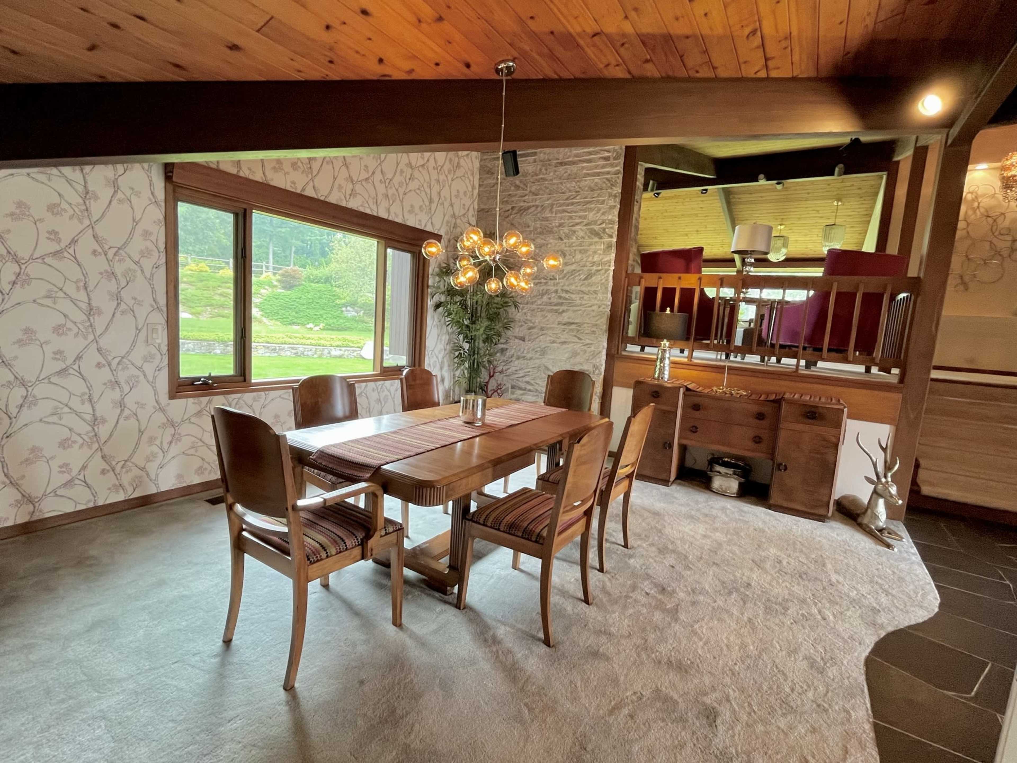 A dining area features a wooden table surrounded by chairs, with a light fixture above and a view of greenery outside through a large window.