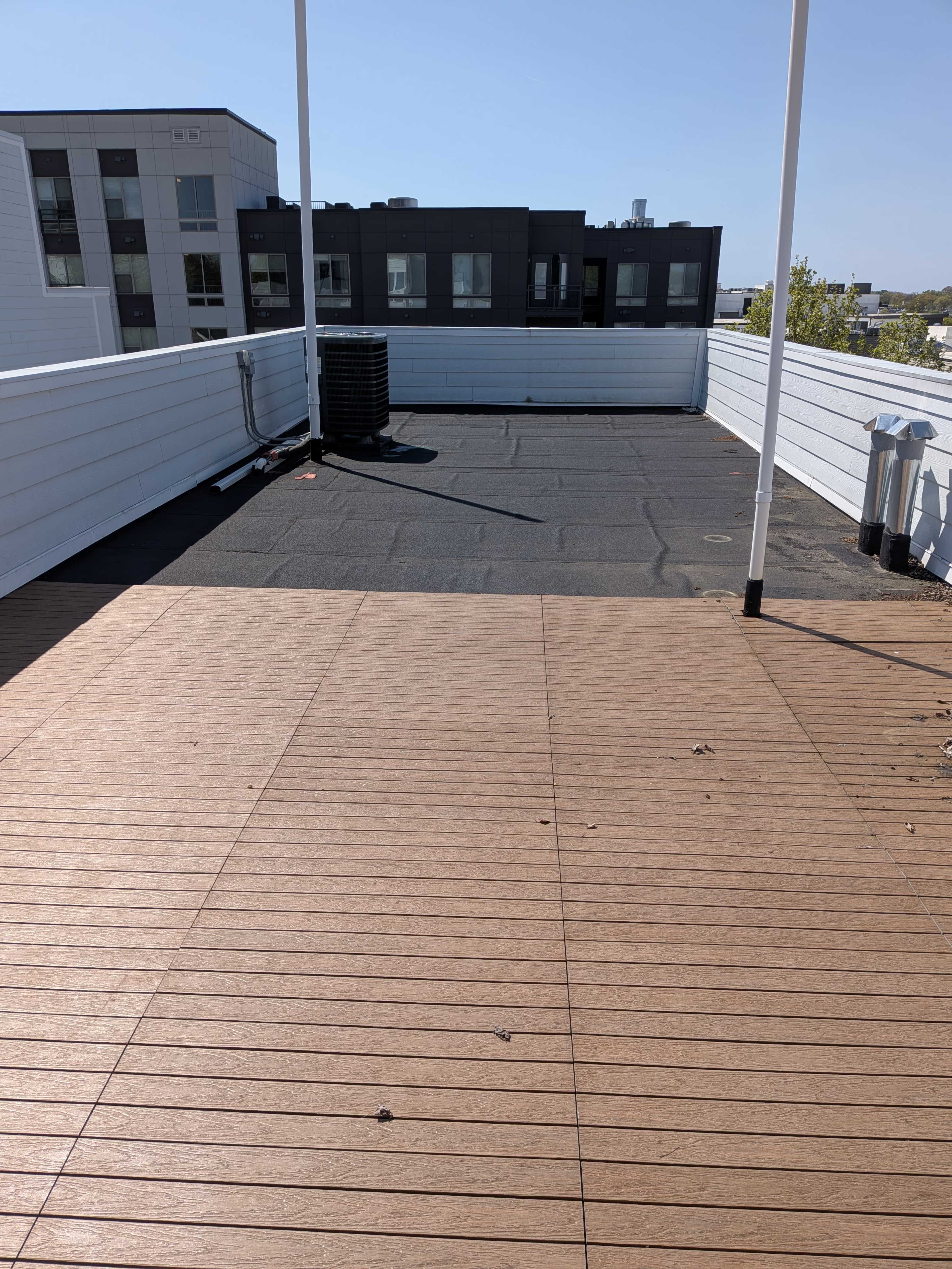 The image shows a flat rooftop terrace with wooden decking, surrounded by a low railing and adjacent buildings in the background.