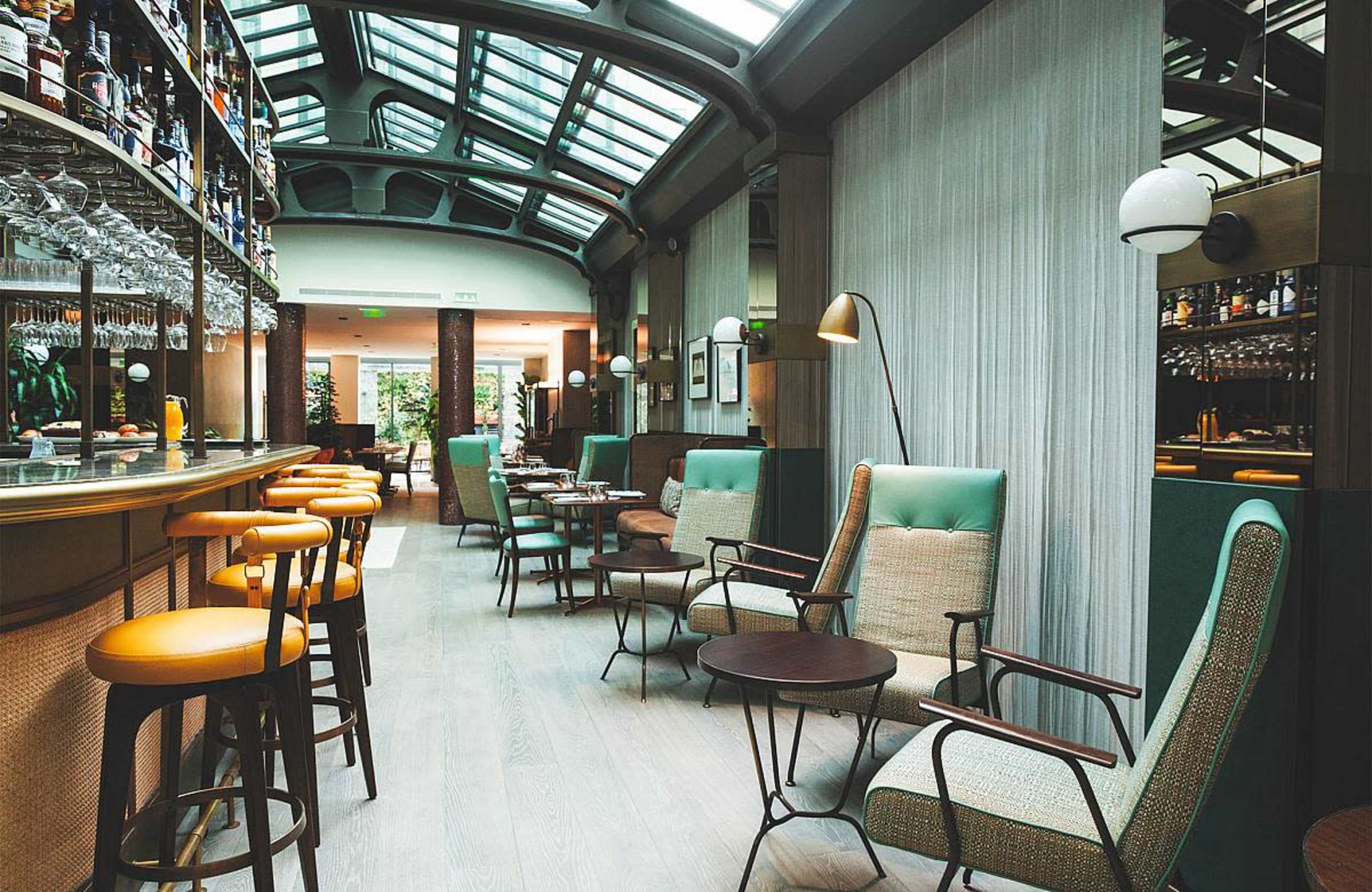 A modern restaurant interior features a bar with high stools, green and beige chairs, and large windows allowing natural light to illuminate the space.