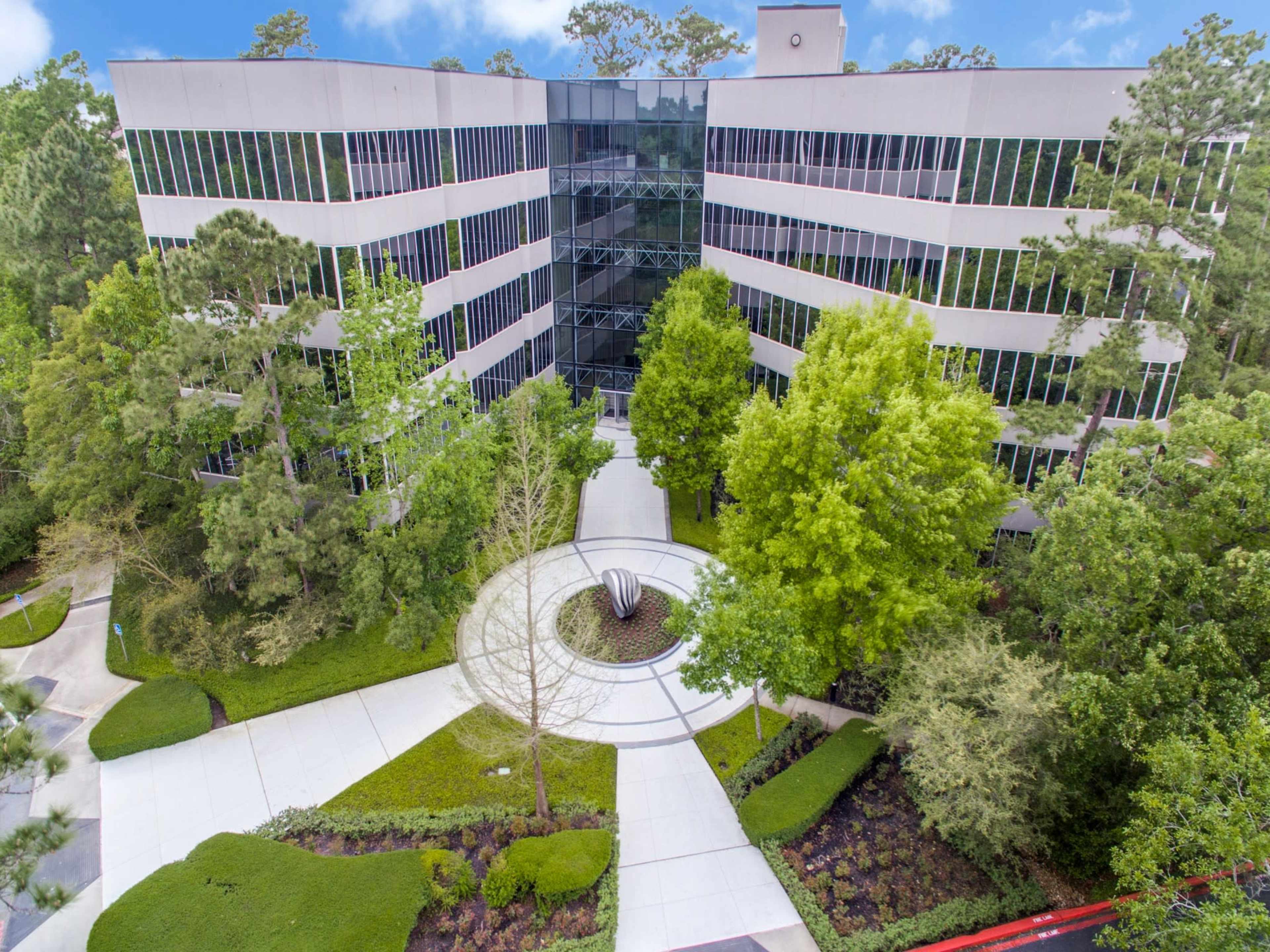 The image shows a multi-story office building surrounded by landscaped greenery and pathways.