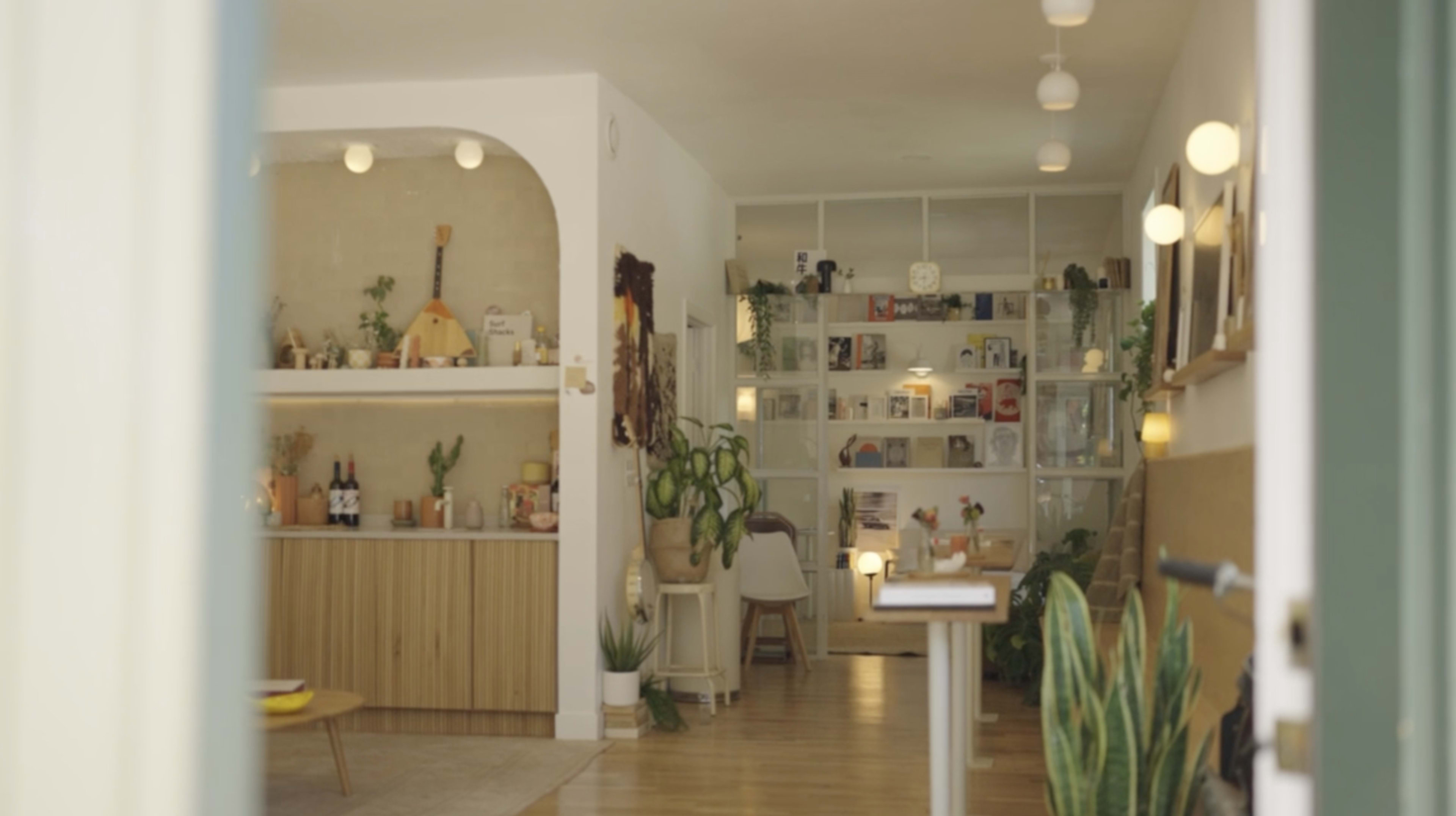 The image shows a bright, modern interior of a cozy café featuring a wooden counter, bookshelves filled with items, and plants decorating the space.