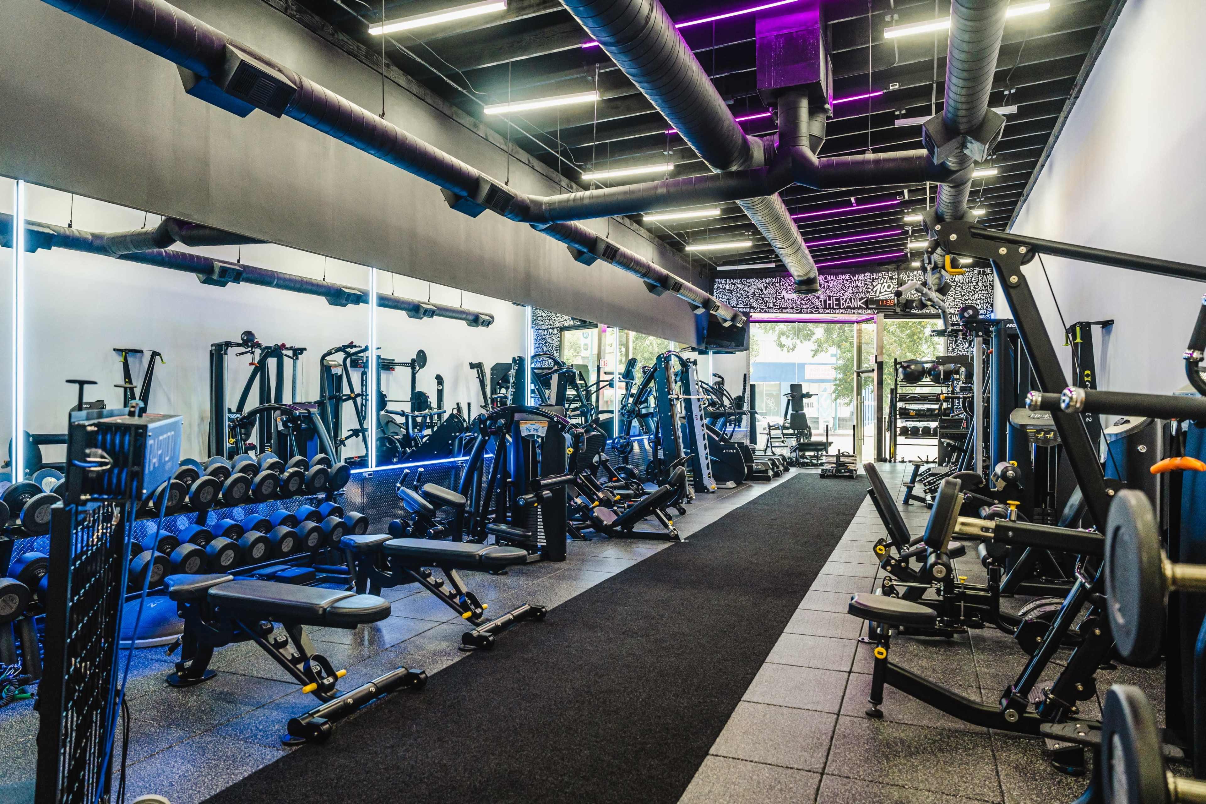 The image shows a modern gym interior with various fitness equipment, including weight machines and free weights, arranged along a long, dark floor.