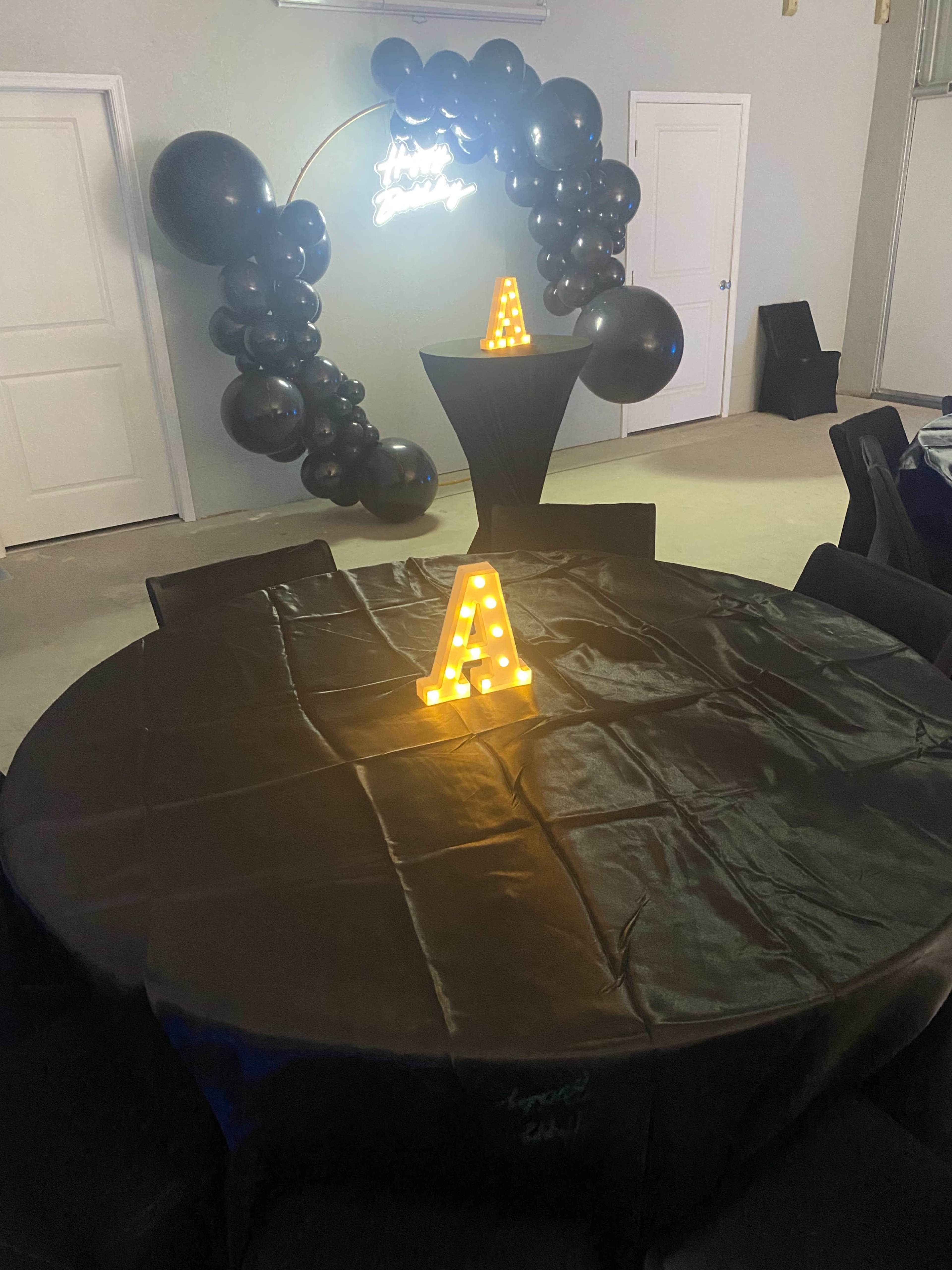 A round table with a dark tablecloth is set up in a room decorated with a black balloon arch and illuminated letter "A" sign.
