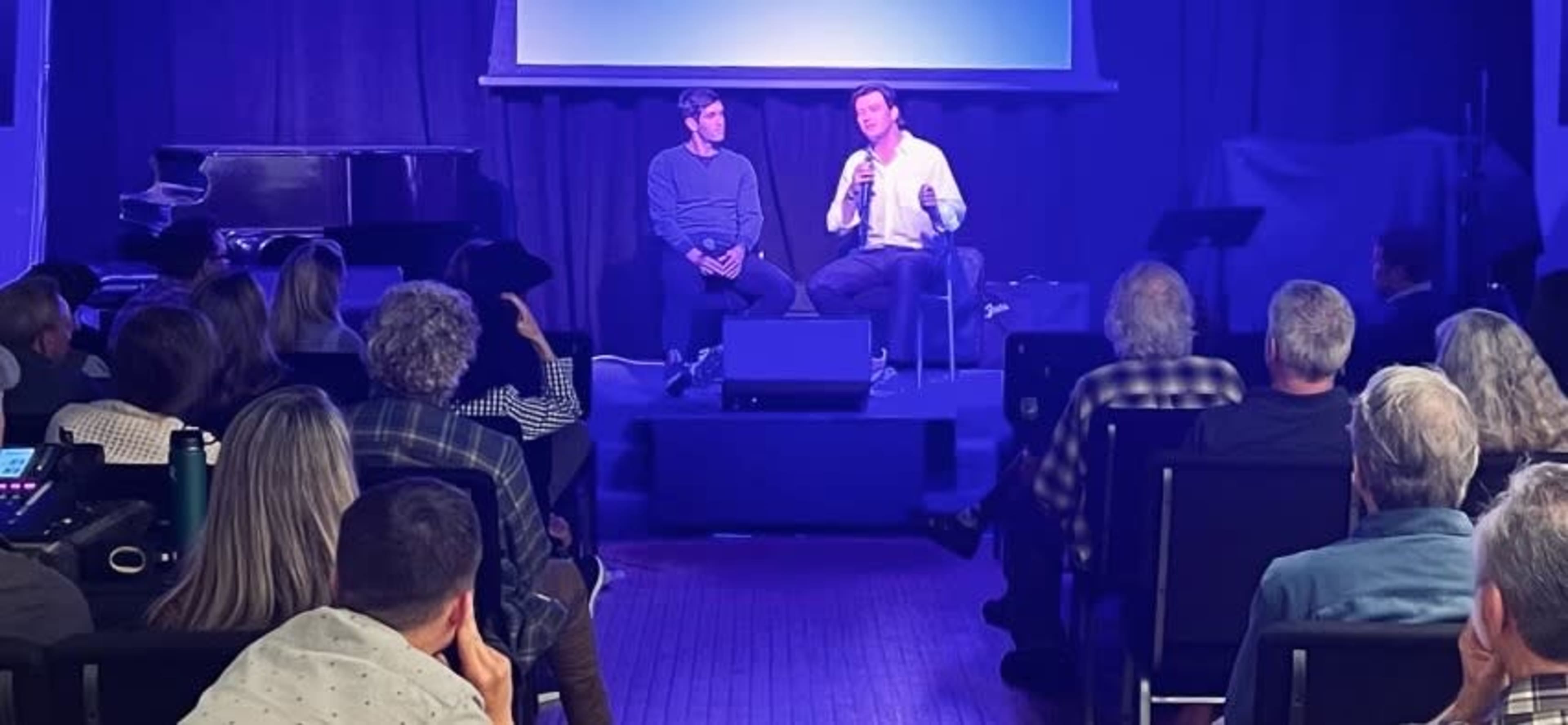 A seated conversation takes place on stage between two speakers in front of an audience, with a piano visible in the background.