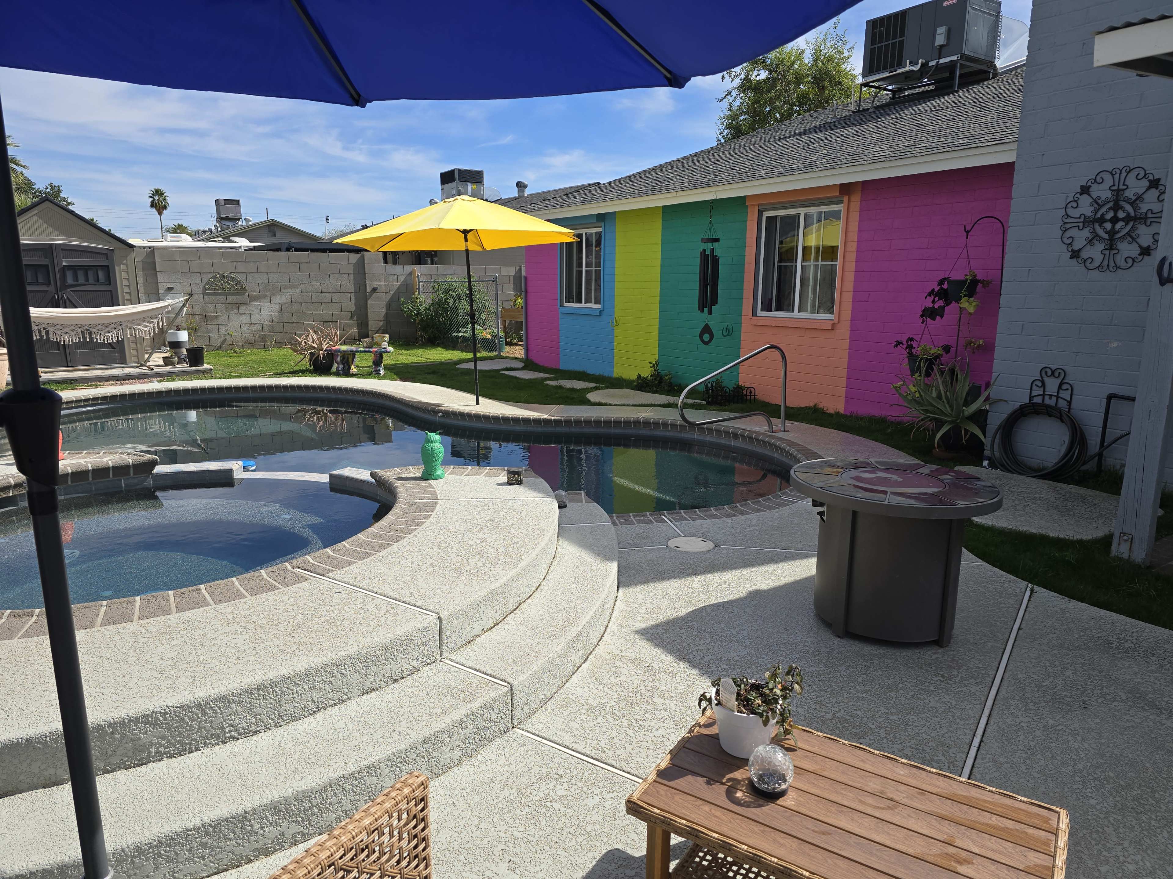The image shows a backyard with a swimming pool, a colorful wall featuring a pink and yellow mural, and various outdoor furniture including an umbrella and a table.
