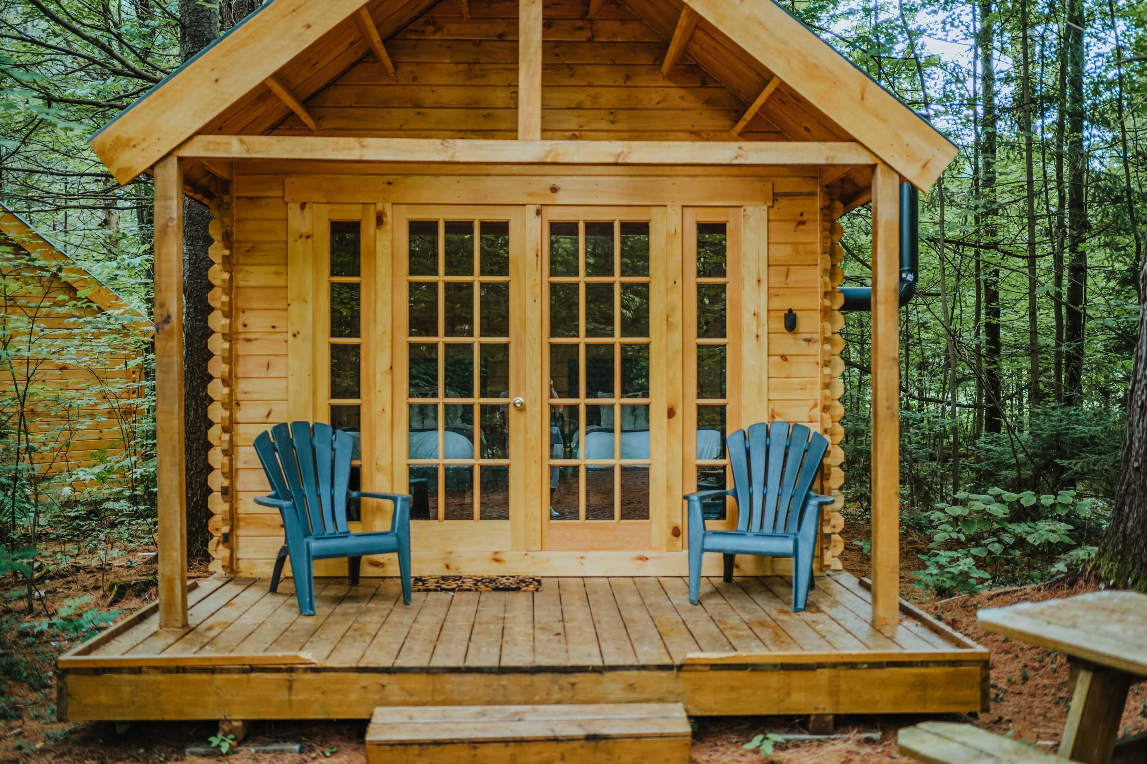 The image shows a rustic wooden cabin with a porch, featuring two blue Adirondack chairs and large glass doors.