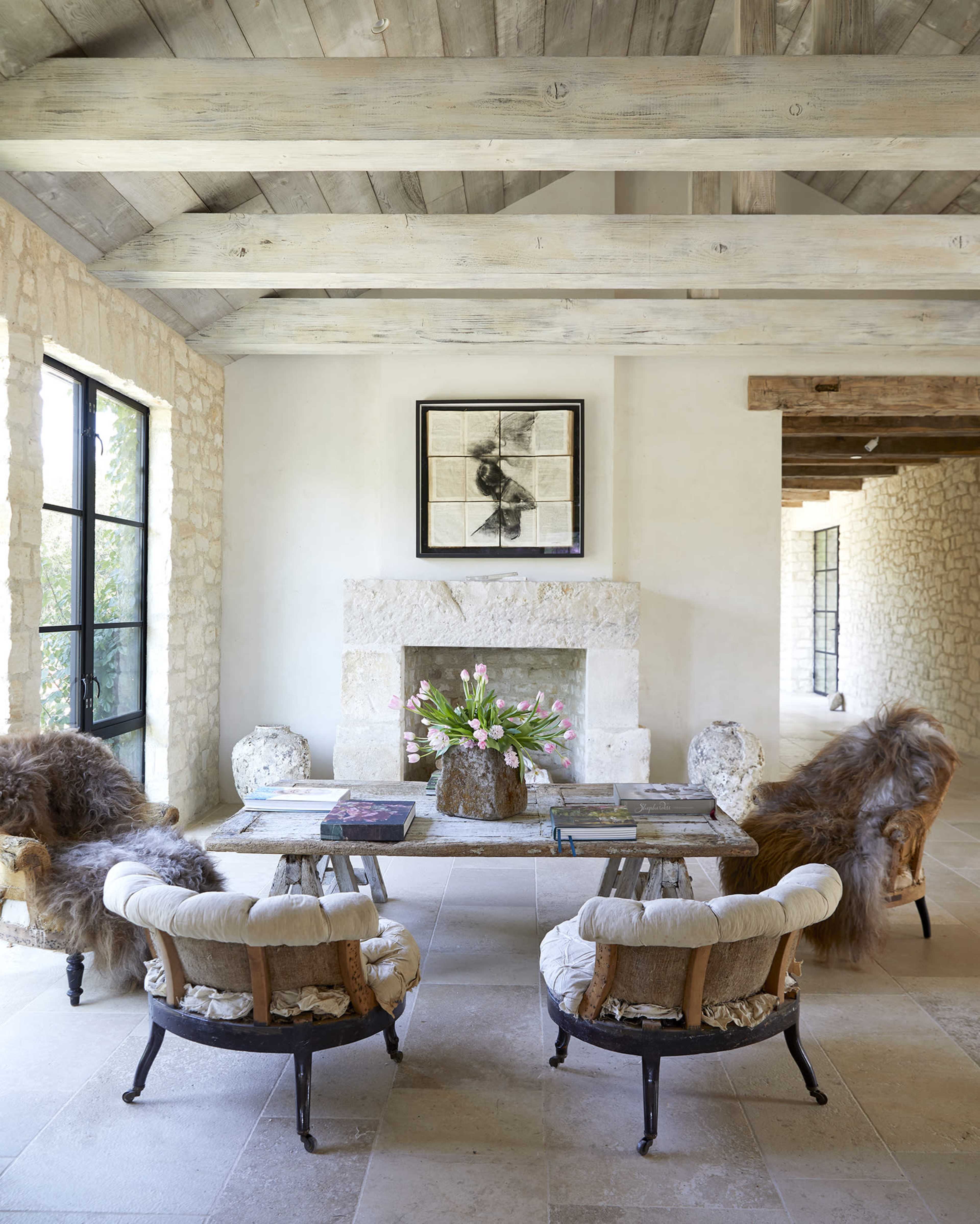 A spacious living area with a stone fireplace, two upholstered chairs, and a table topped with magazines and a vase of flowers.