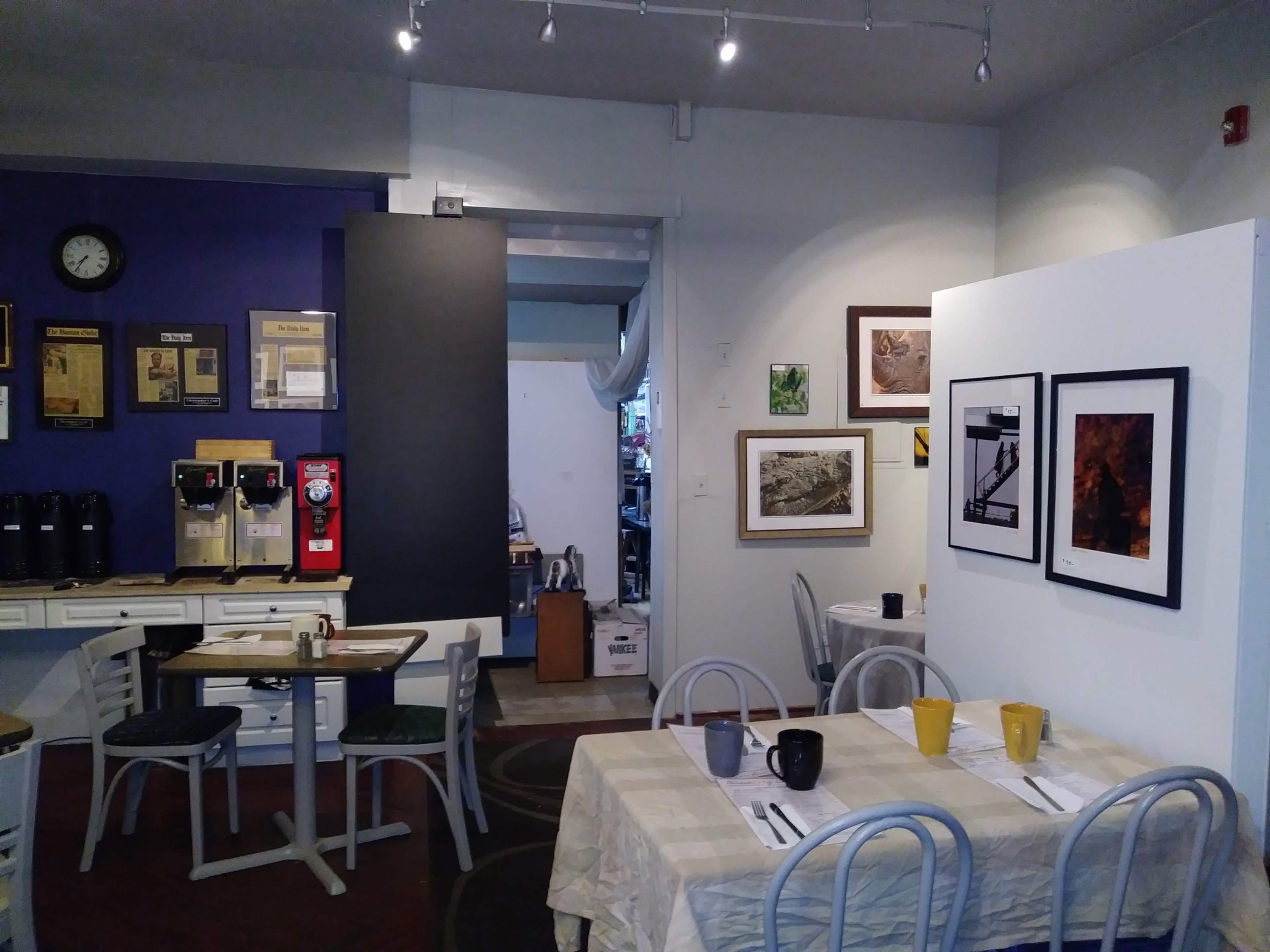 The image shows a cozy café interior with tables set for dining, artworks displayed on the walls, and a counter area featuring coffee dispensers and informational materials.